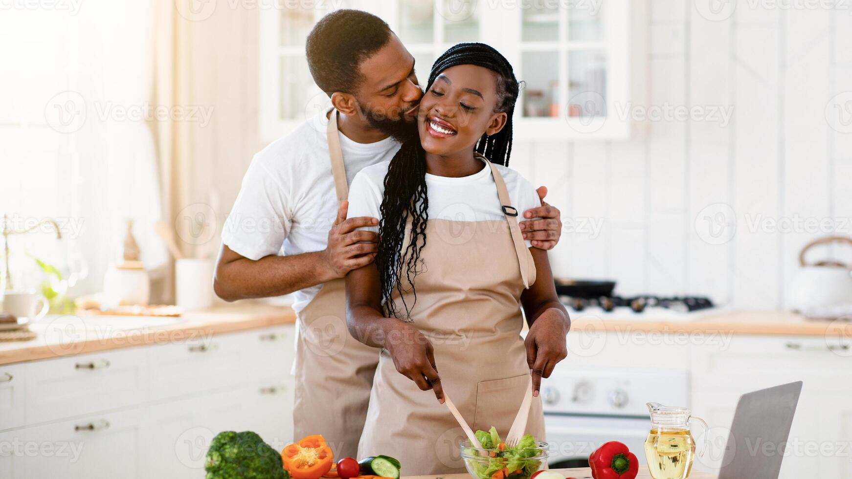 Romantic Black Couple Kissing While Cooking Healthy Food Together In Kitchen. Affectionate African American Spouses Preparring Vegetarian Lunch On Table With Laptop, Enjoying Time Together, Free Space photo
