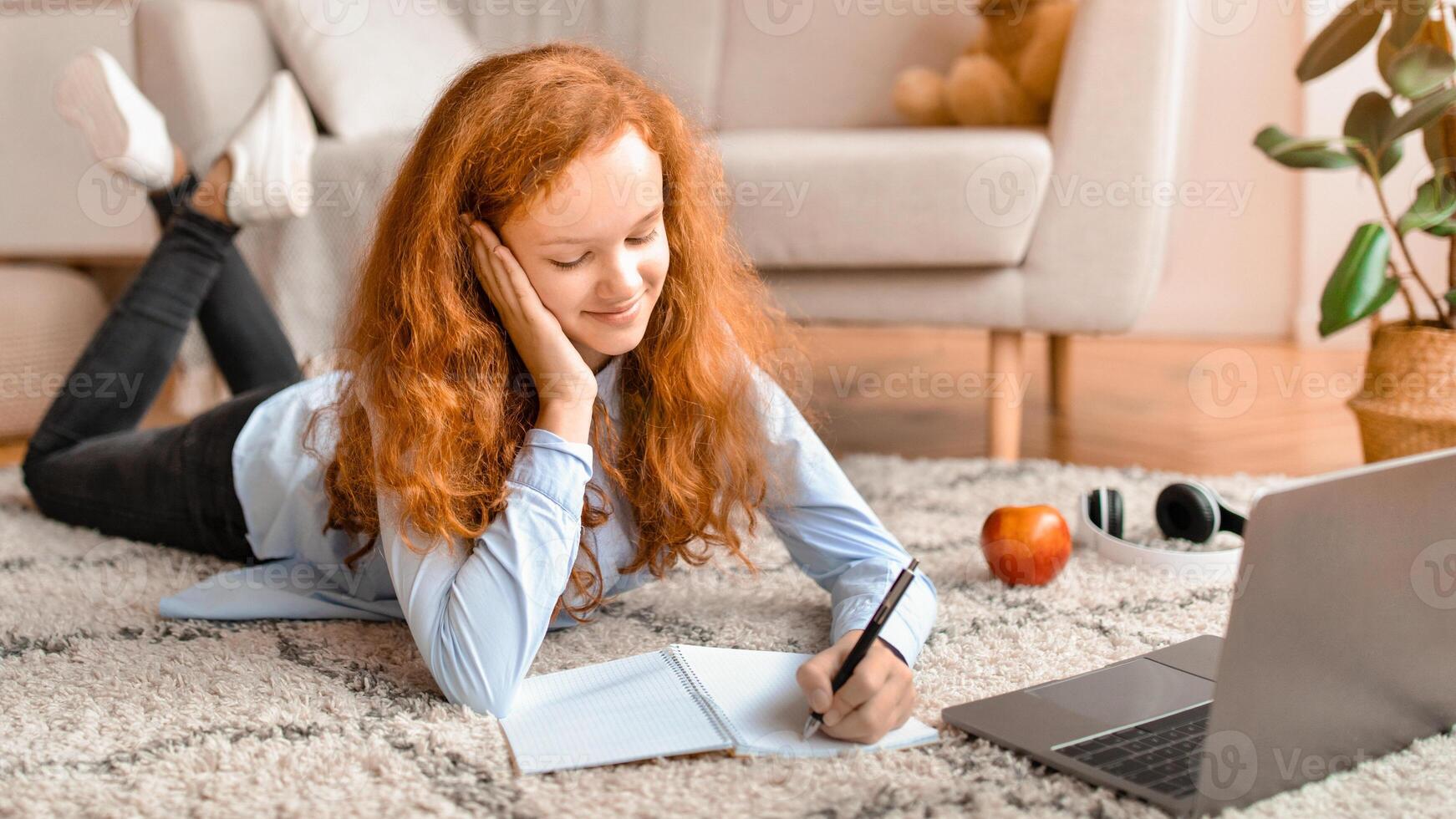 Distance Education. Smiling schoolgirl lying on the floor carpet with her laptop and writing in notebook. Girl watching tutorial, lecture or webinar, studying online at home. E-learning photo