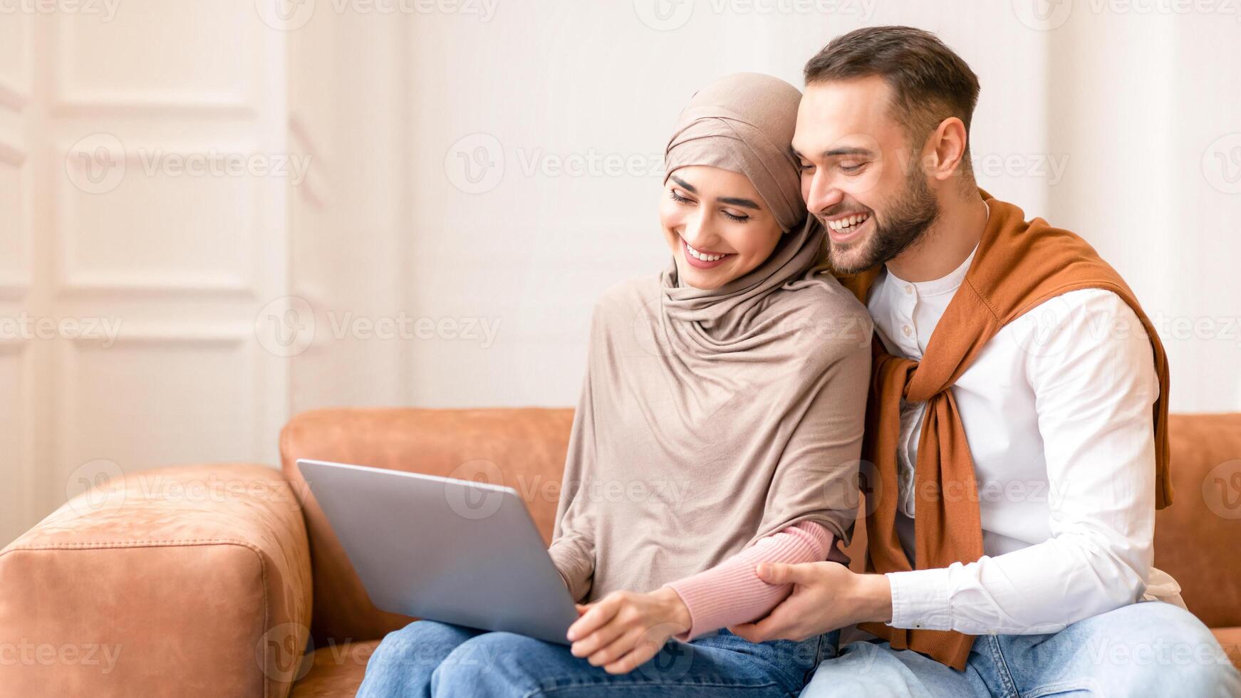 Cheerful Married Muslim Couple Using Laptop Computer Together Sitting On Sofa At Home. Modern Arab Family Watching Movie Online Relaxing On Weekend. People And Technology, Internet Concept photo