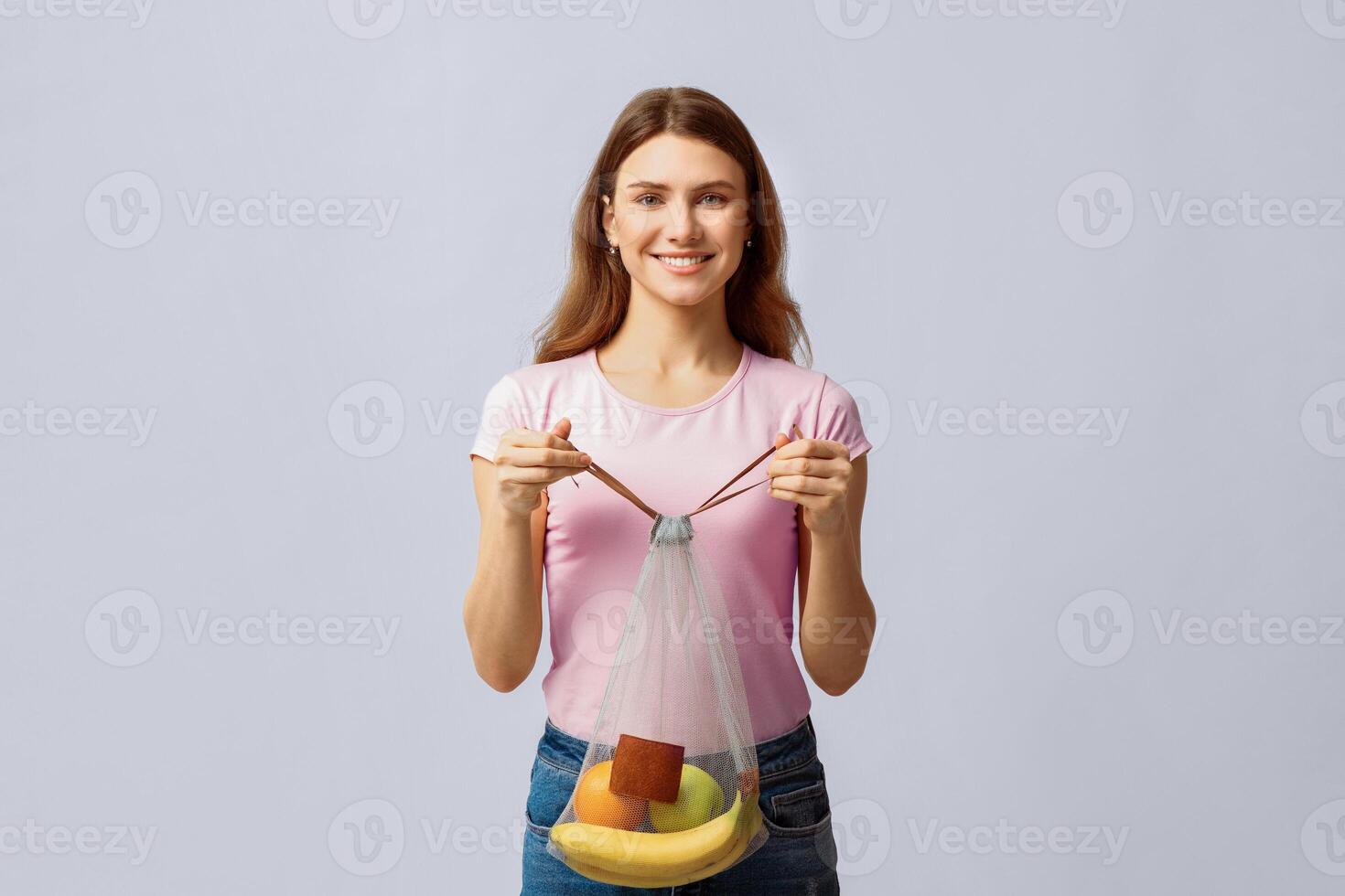 A young woman holds a reusable mesh bag filled with fruits, promoting sustainable living and eco-friendly habits. Her cheerful demeanor suggests a commitment to environmental awareness. photo