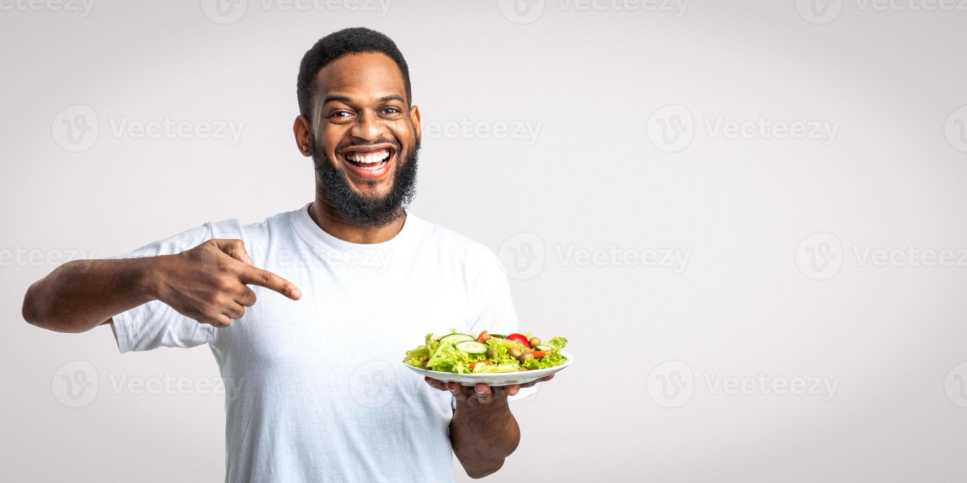 Eat Healthy. Cheerful Black Guy Pointing Finger At Plate With Vegetable Salad Dieting Standing Over White Studio Background. Male Nutrition And Weight Loss, Slimming Concept photo
