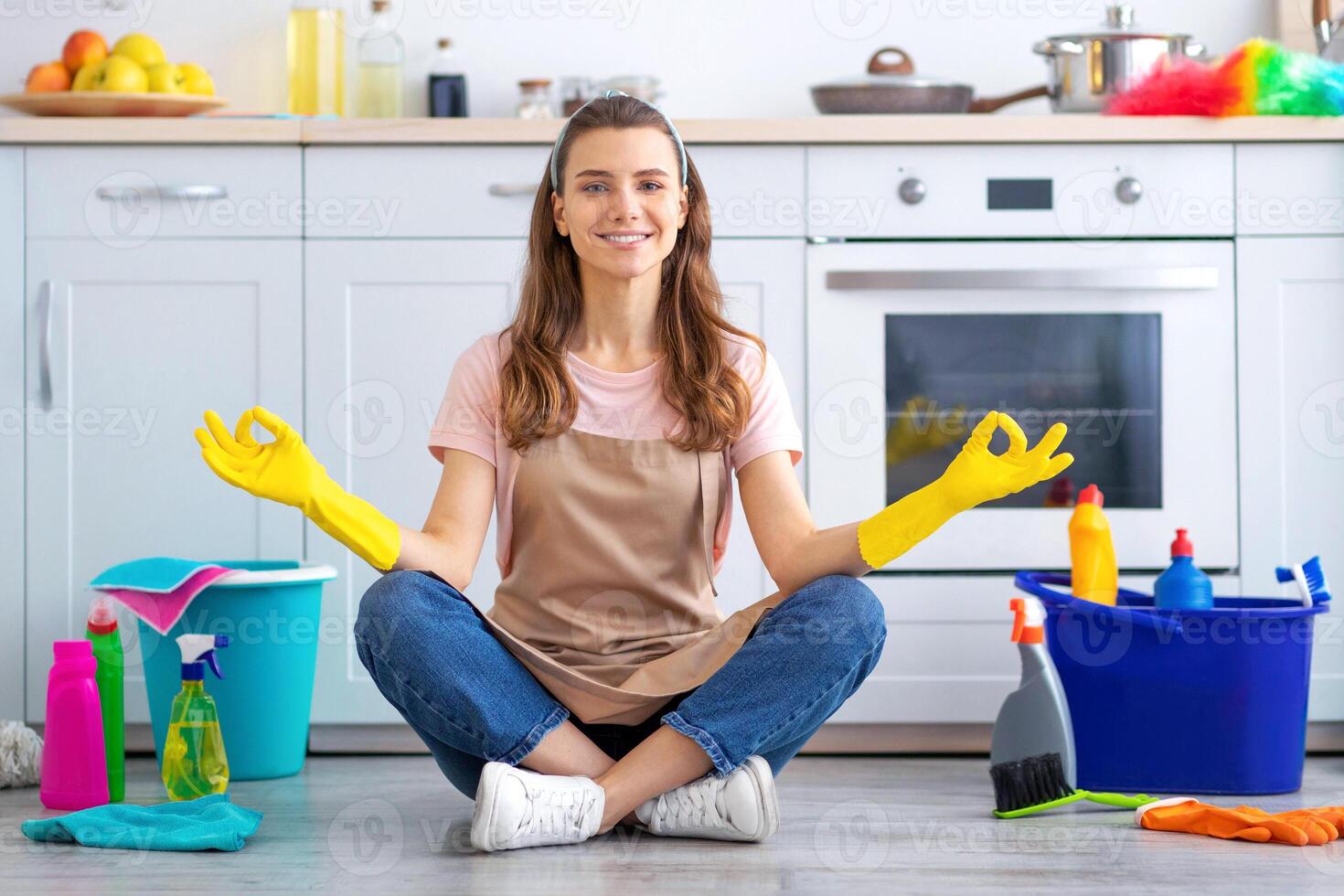 Happy millennial housewife sitting in lotus yoga pose on kitchen floor surrounded by cleaning supplies. Balanced young maid taking break from domestic duties, meditating during home cleanup photo