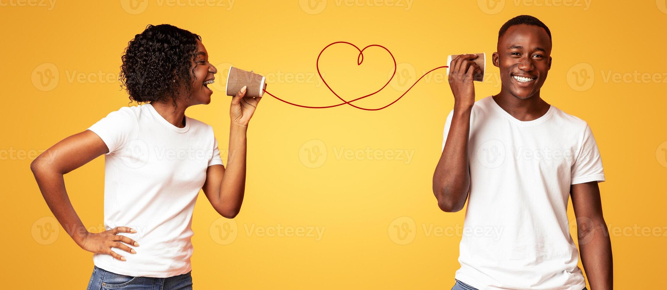 Communication and fun during Valentine Day. Young black couple talking through tin can phone, orange studio background. African american lady saying words of love to her boyfriend photo
