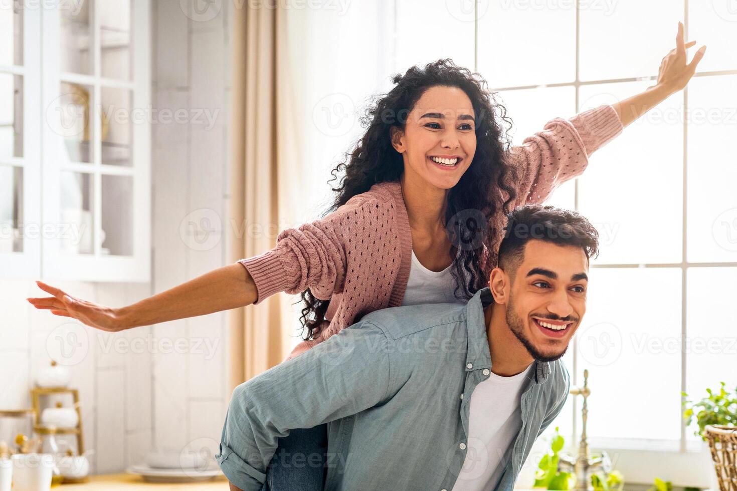 Joyful Middle Eastern Spouses Having Fun Together In Kitchen Interior, Handsome Arab Man Piggybacking His Cheerful Wife, Happy Attractive Lady Enjoying Ride, Spreading Hands Like Flying, Copy Space photo