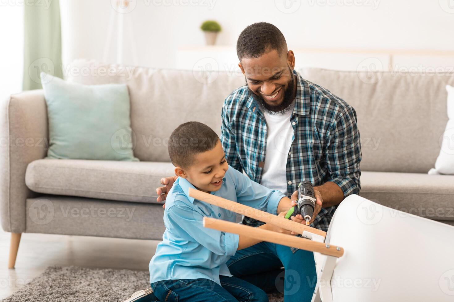 African American Father And Son Fixing Table Together Using Electric Drill Doing House Chores At Home. Happy Kid Helps Dad With Housework Concept. Selective Focus photo
