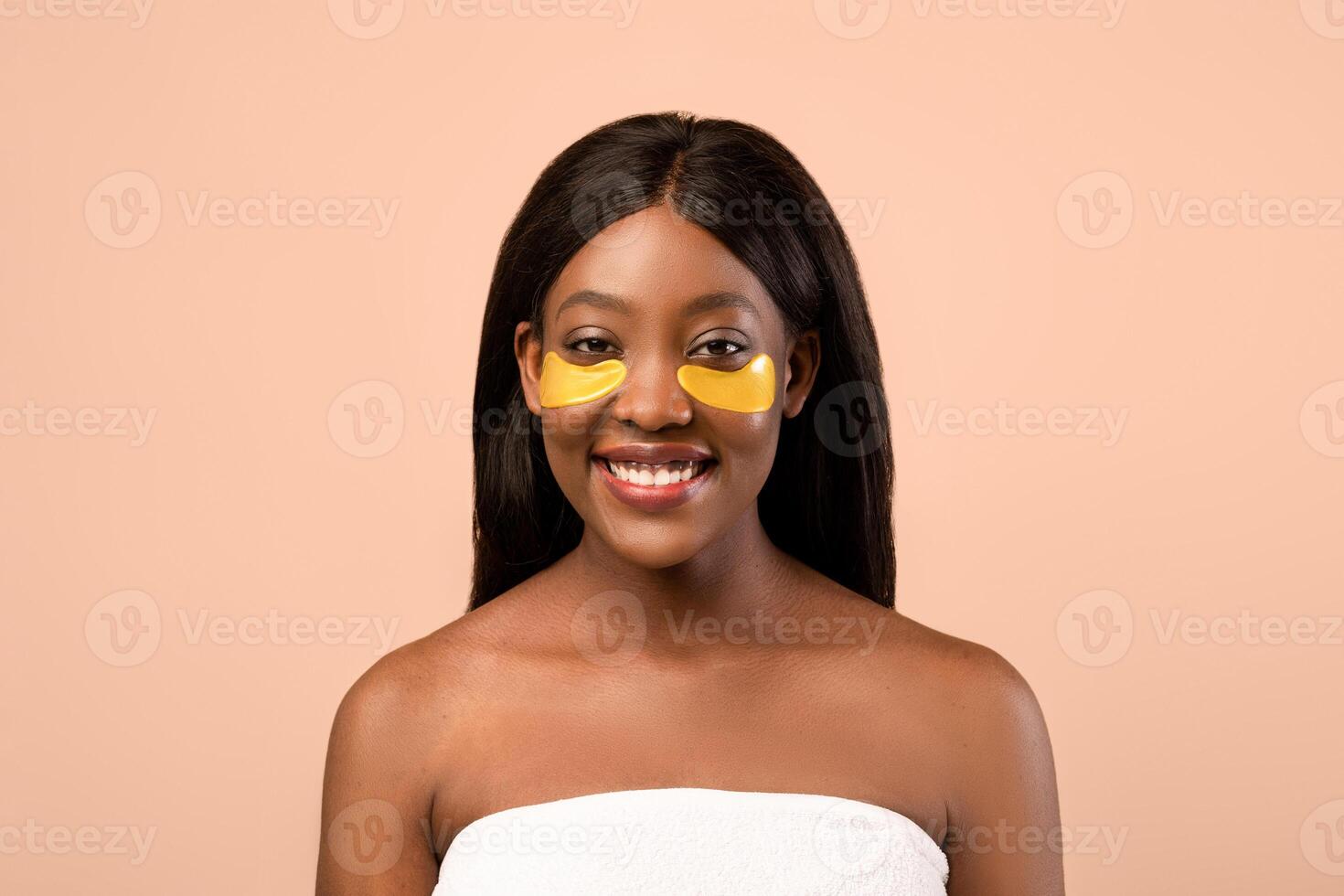 Cheerful attractive young black woman wrapped in towel using golden hydrogel eye patches over beige studio background, copy space. Smiling fair-haired african american lady making eyes care photo