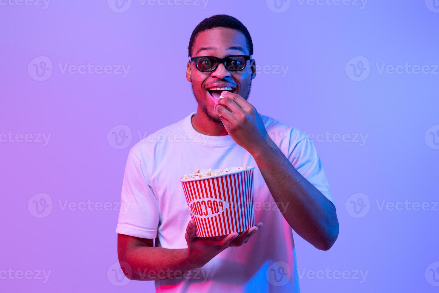 Cinema Time. Excited Black Man In 3d Glasses Eating Popcorn And Enjoying Movie, Looking At Camera With Amazement, Cheerful African American Guy Standing In Vivid Neon Light On Purple Background photo