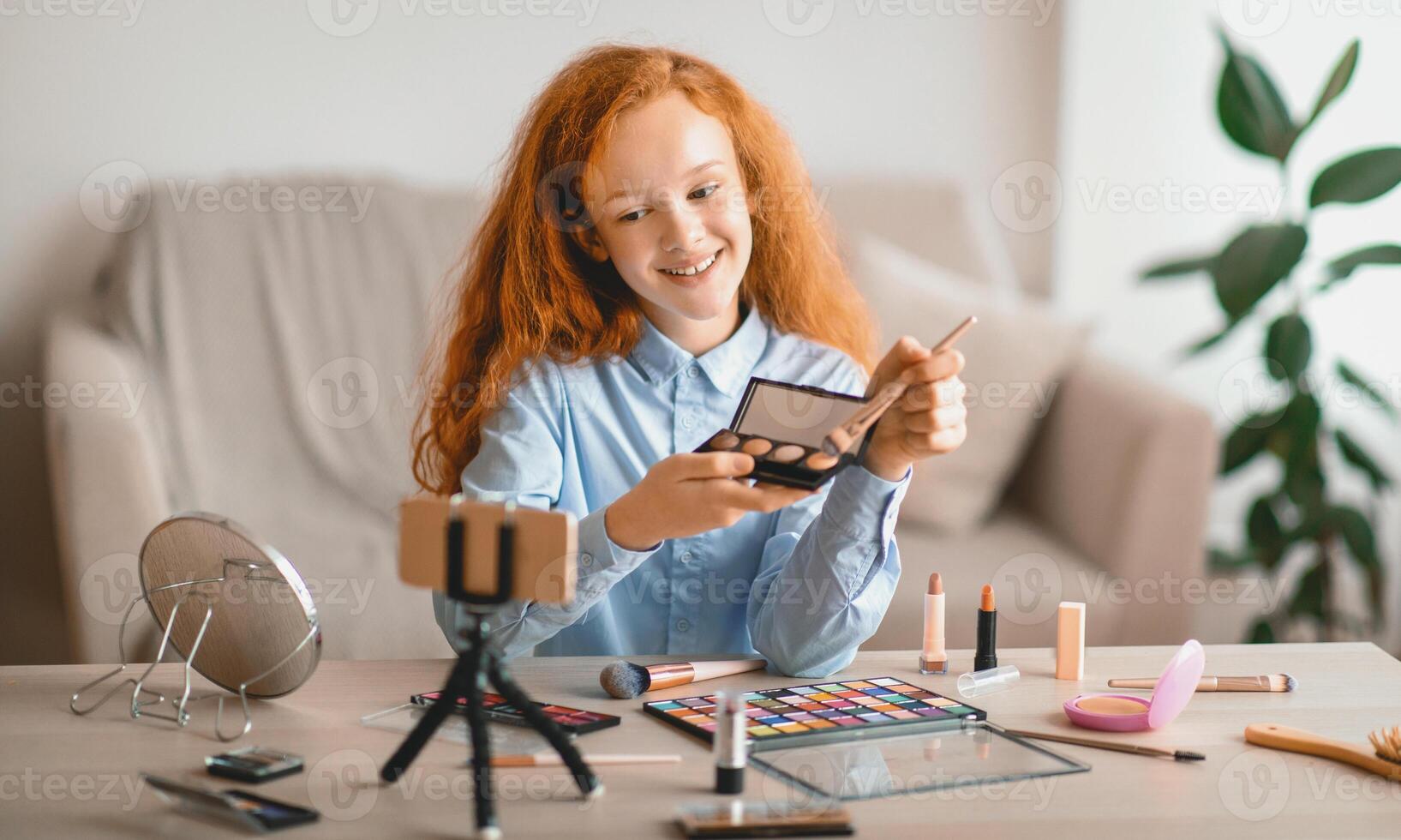 Live Streaming And Blogging Concept. Portrait of smiling teenage girl doing make up, showing her eyeshadow palette, looking at camera, shooting and filming vlog using cellphone on tripod photo
