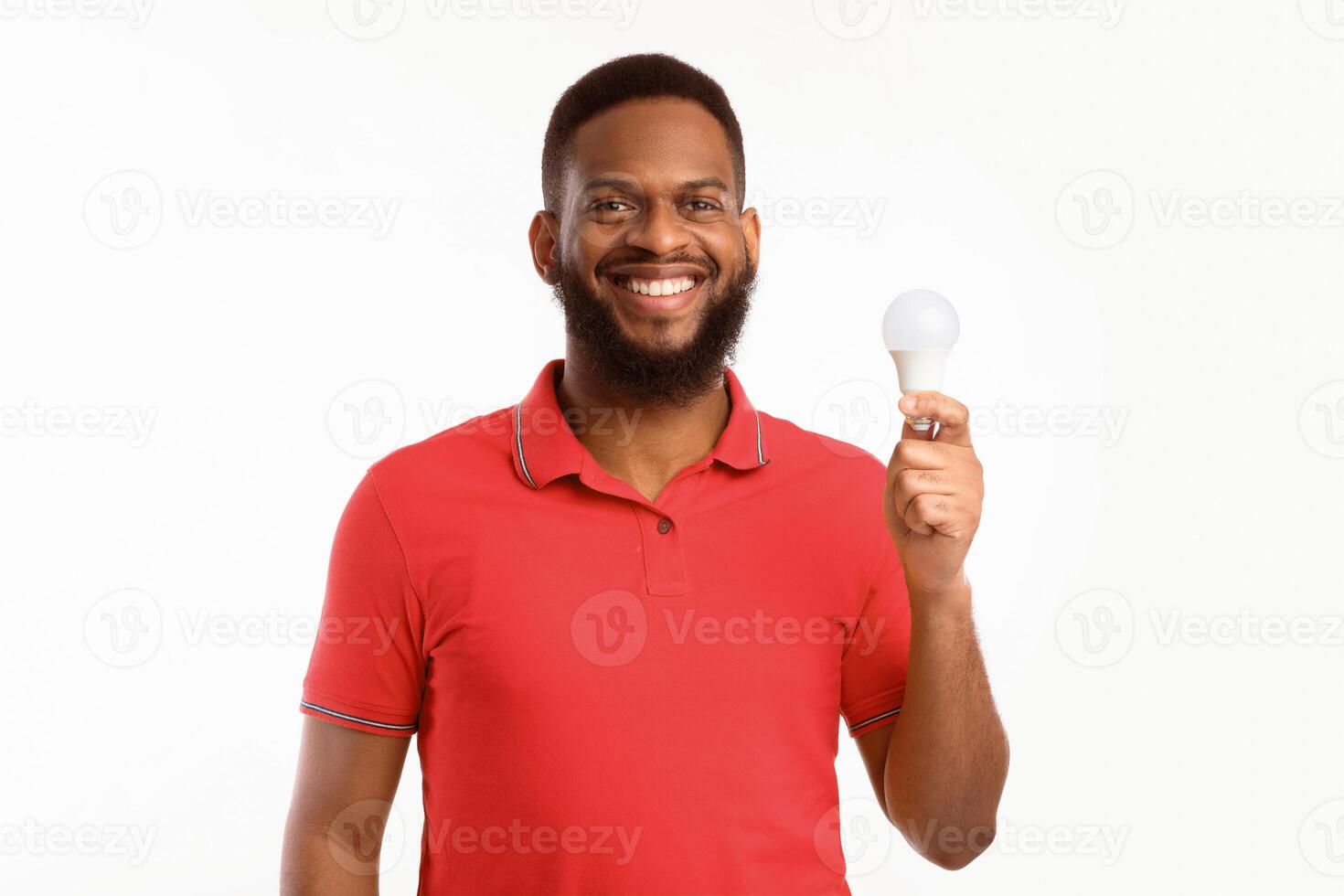 Happy Black Guy Showing Energy-Saving Lightbulb Standing Over White Studio Background, Posing Smiling To Camera. Save Energy, Light Bulb Disposal Service Concept photo