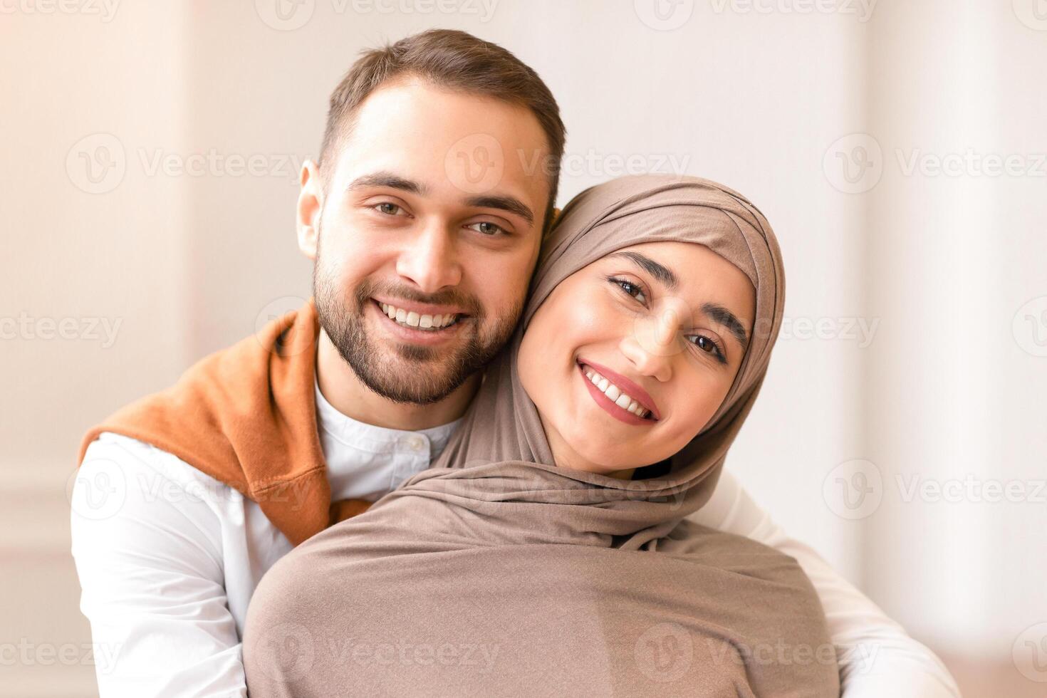 Cheerful Young Muslim Couple Hugging Posing Smiling To Camera Standing Indoors At Home. Arab Spouses In Love. Happy Arabic Family Portrait. Happiness In Marriage And Romantic Relationship Concept photo