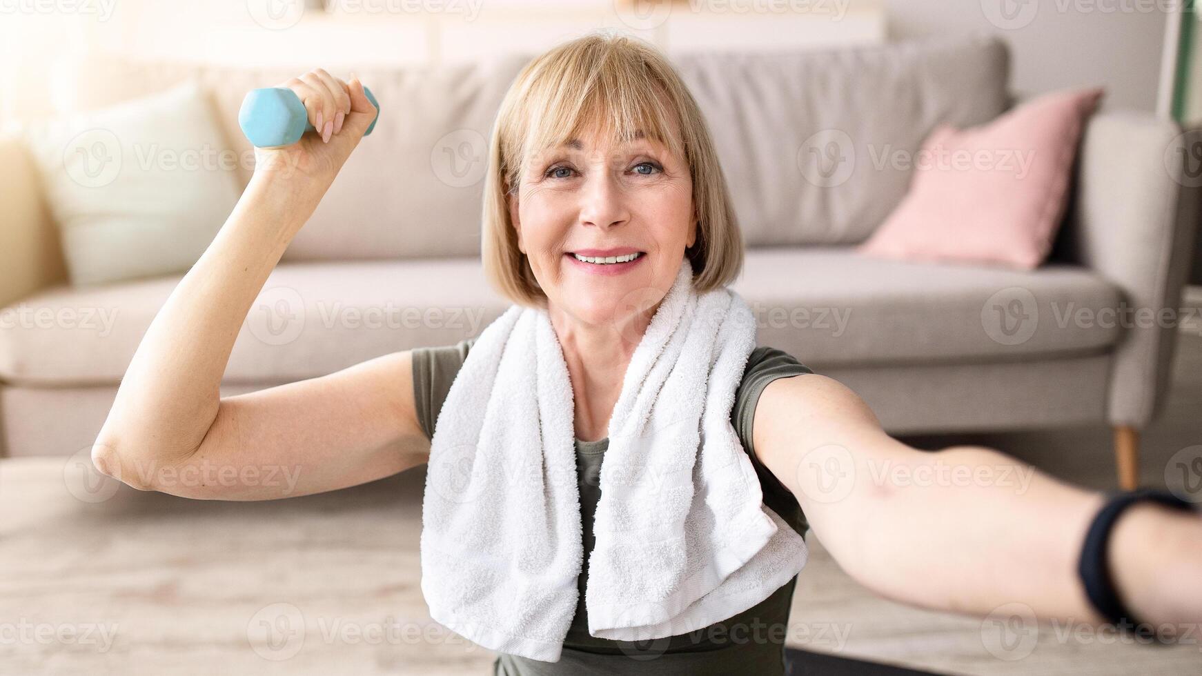 Joyful mature woman taking selfie while exercising with dumbbells at home. Happy senior lady making photo of herself during domestic strength workout, enjoying her training