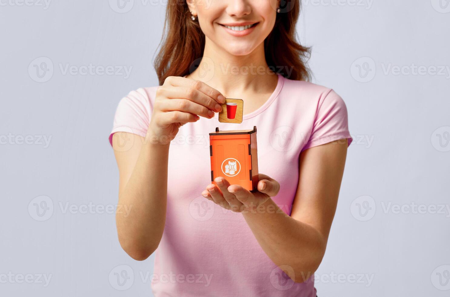 Unrecognizable Lady Putting Card With Plastic Trash Icon Into Little Orange Garbage Bin, Smiling Woman Sorting Waste For Recycling, Standing Over Grey Background In Studio, Cropped Image, Free Space photo