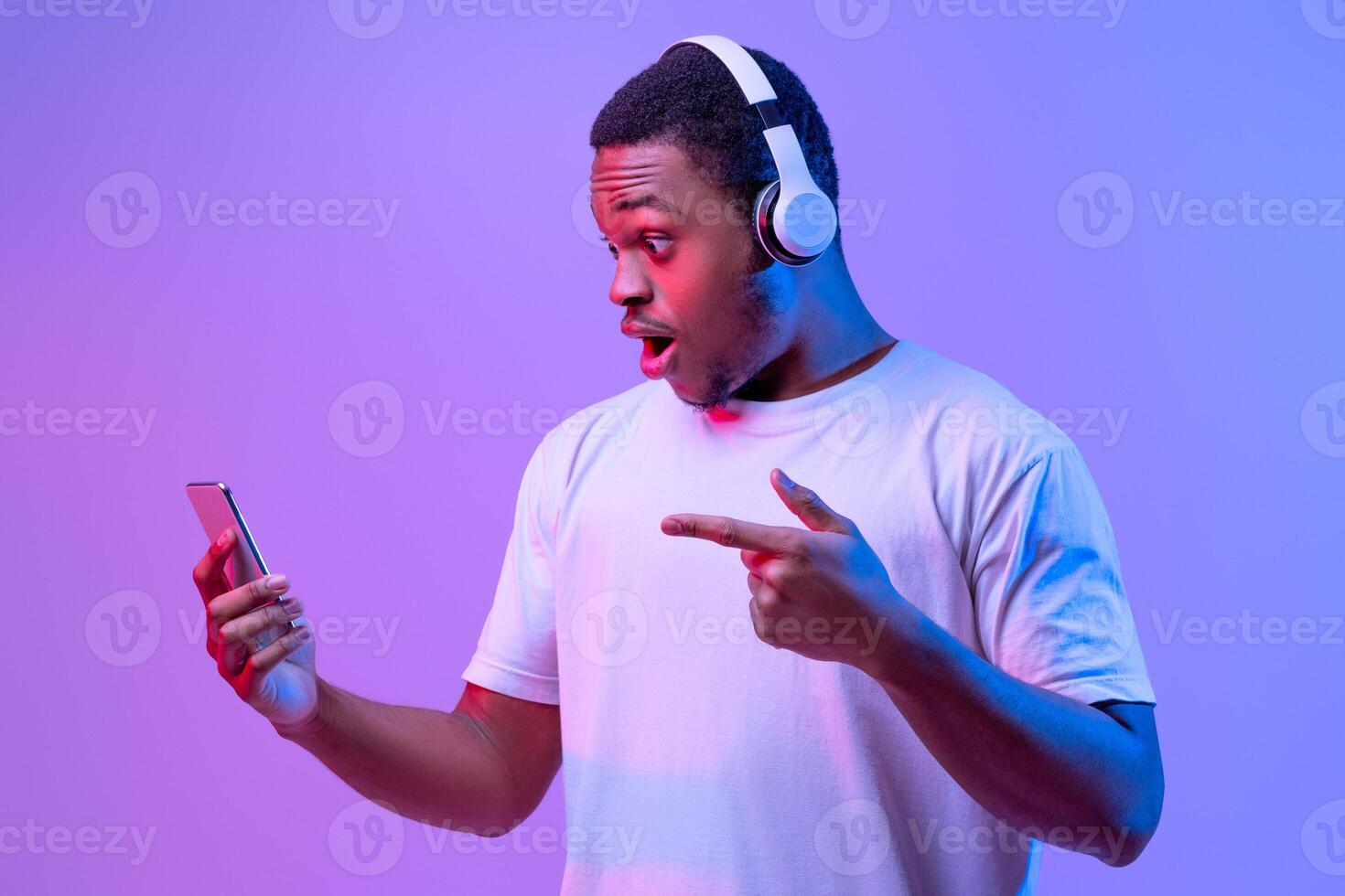 Amazed Black Guy In Wireless Headphones Pointing At Smartphone In His Hands, Excited African American Man Emotionally Reacting To Mobile Offer, Standing In Neon Lighting Over Purple Background photo
