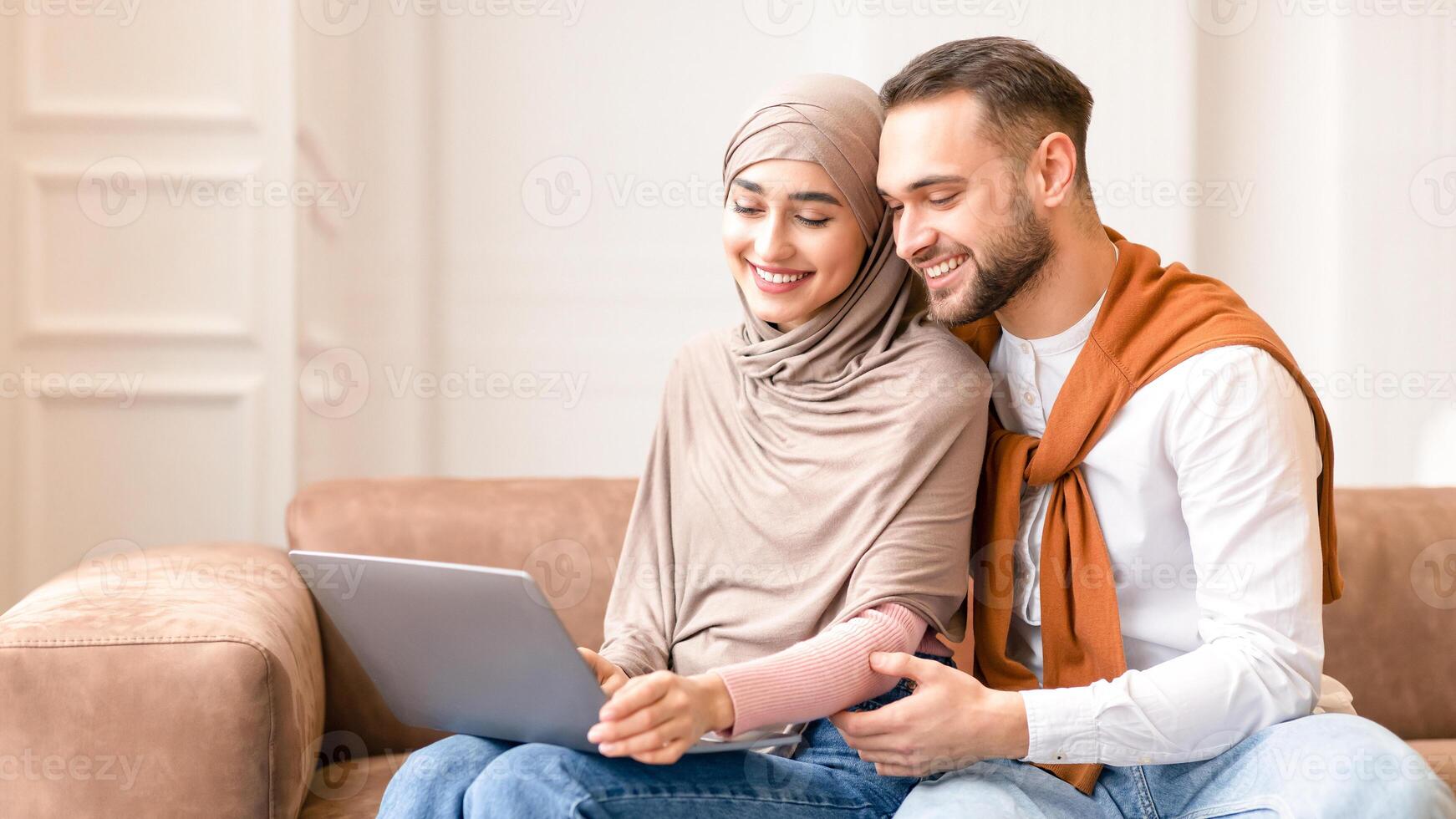 Happy Married Arab Couple Watching Movie On Laptop Sitting And Hugging On Sofa In Living Room At Home. Weekend Leisure And Relaxation. Muslim Spouses Browsing Internet Together photo