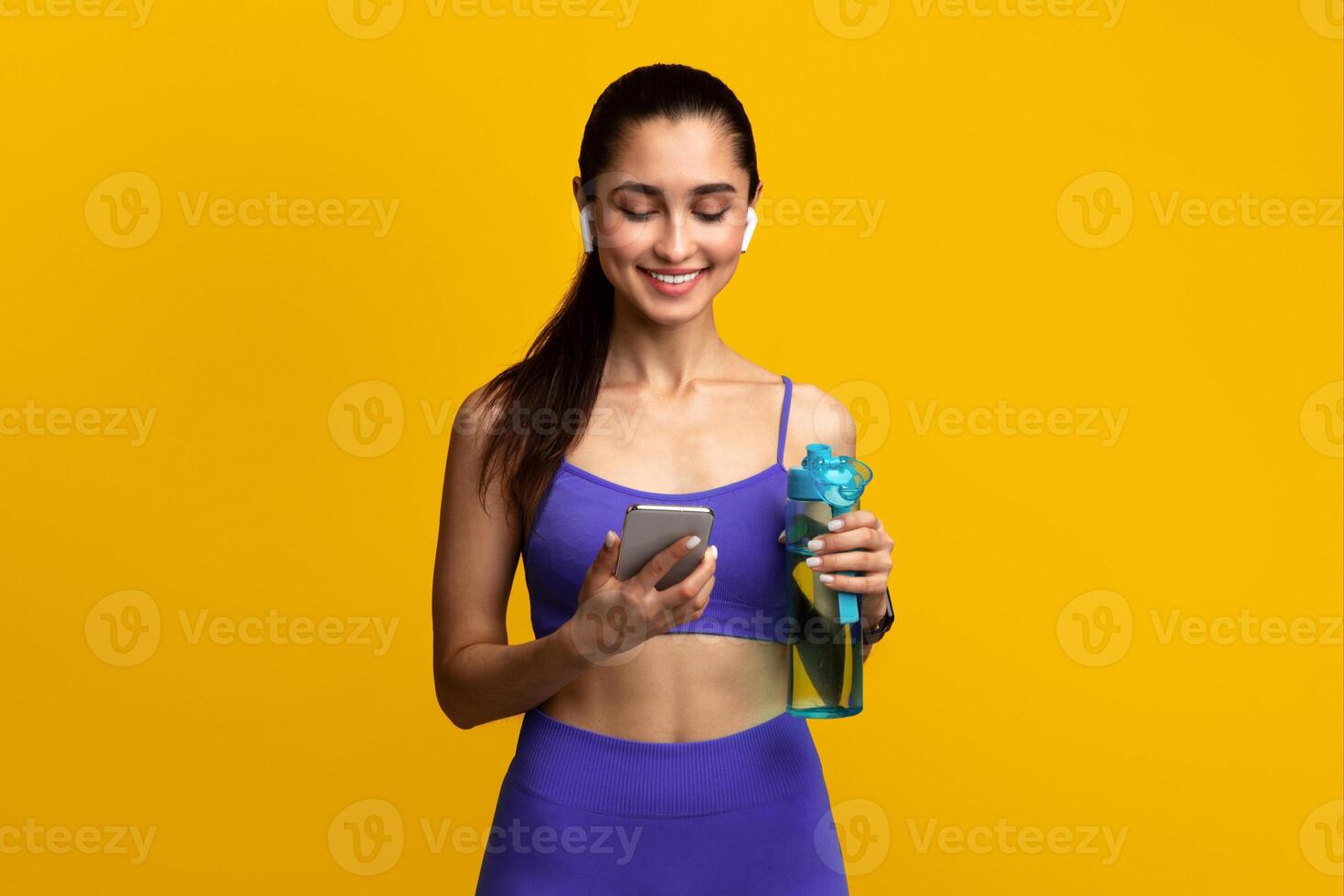 Music And Sport. Portrait of happy smiling woman in sportswear and wireless earphones holding bottle with fresh water and using mobile phone, standing isolated on orange studio background photo