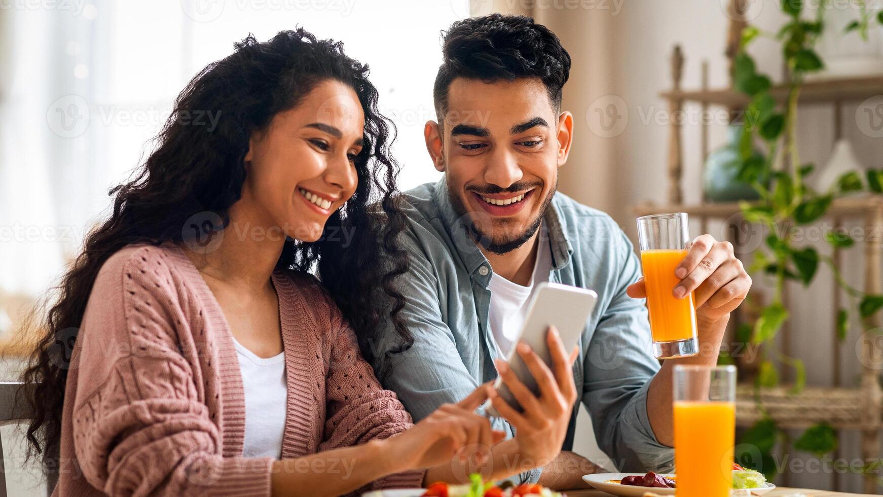 Happy middle eastern couple watching photos on smartphone while having breakfast in kitchen, young arab spouses spending time with cellphone and enjoying tasty morning meal, closeup shot