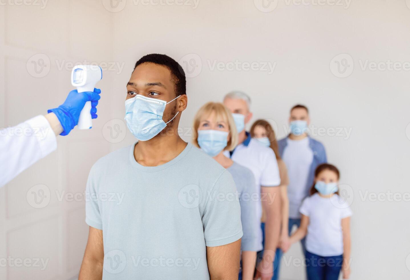 Coronavirus immunization medical campaign concept. Black man in face mask standing in line at clinic, doctor measuring his temperature with infrared thermometer before covid-19 vaccination, copy space photo