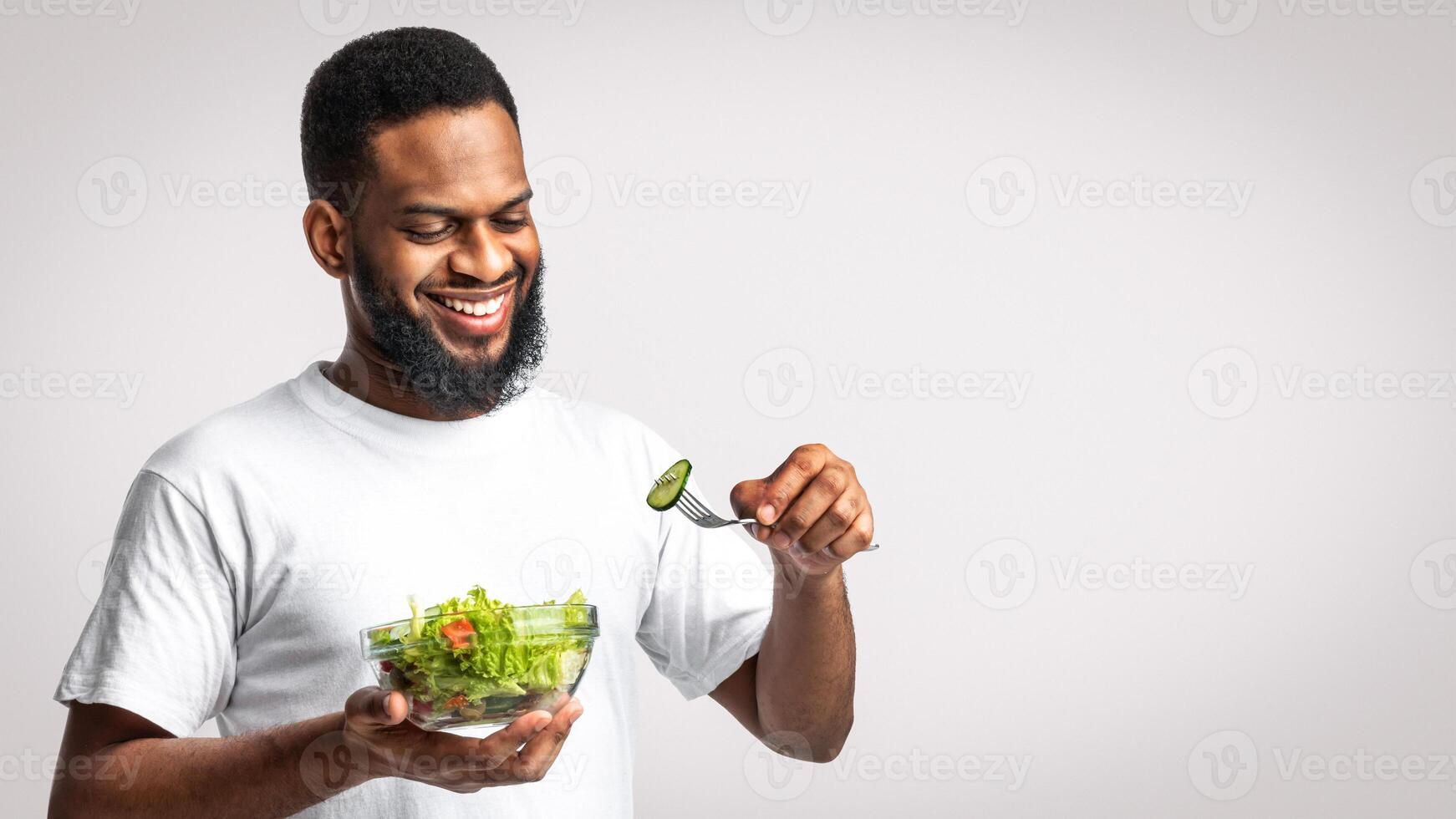 Eat Healthy. Happy African American Man Eating Fresh Vegetable Salad Standing Posing With Plate Over White Studio Background. Dieting, Nutrition And Male Weight Loss Concept photo