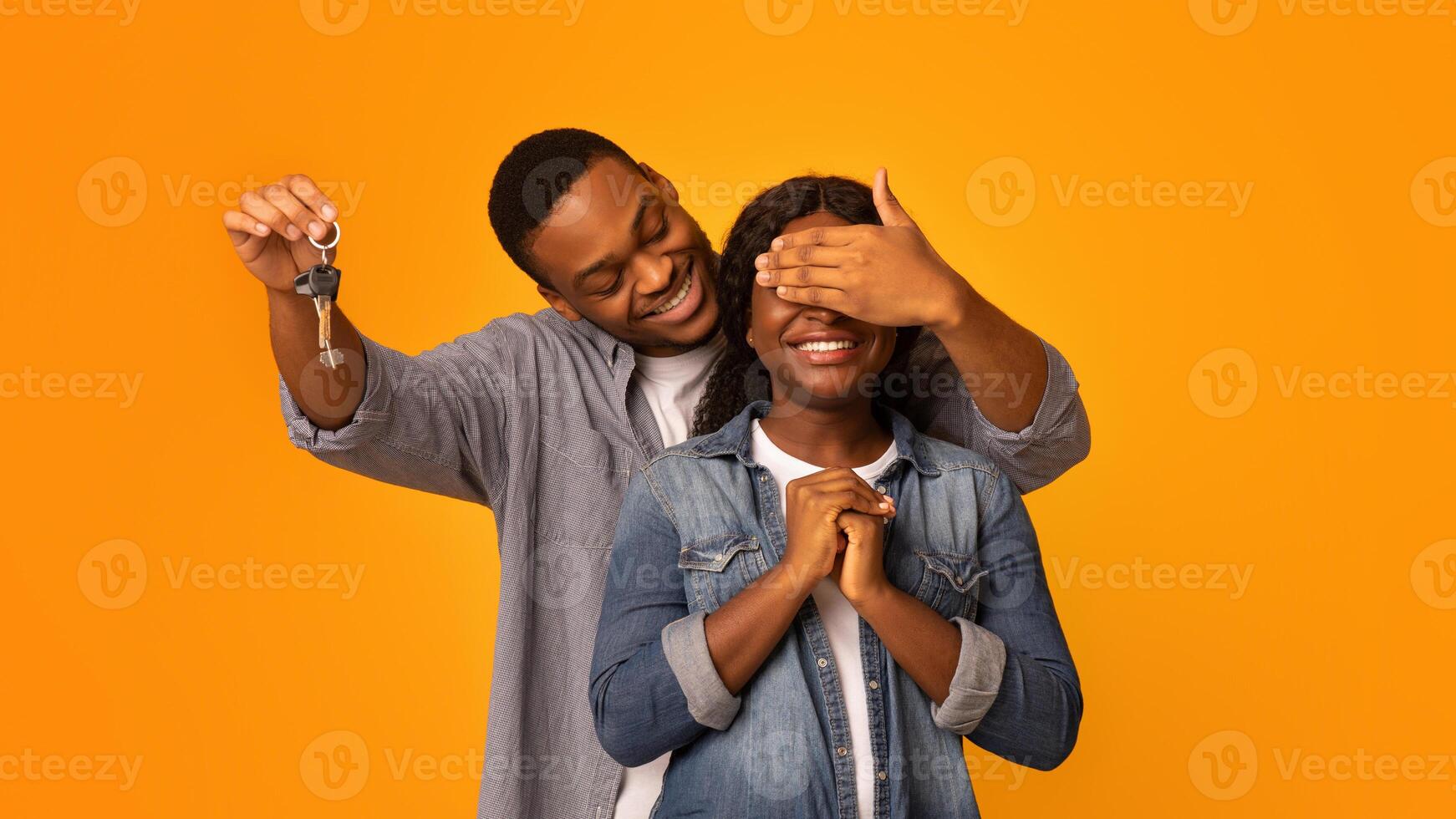 New Home. Loving Black Husband Holding Keys And Covering Wife's Eyes, Surprising His Excited Woman With Buying New Property While Standing Together Isolated Over Yellow Background, Copy Space photo