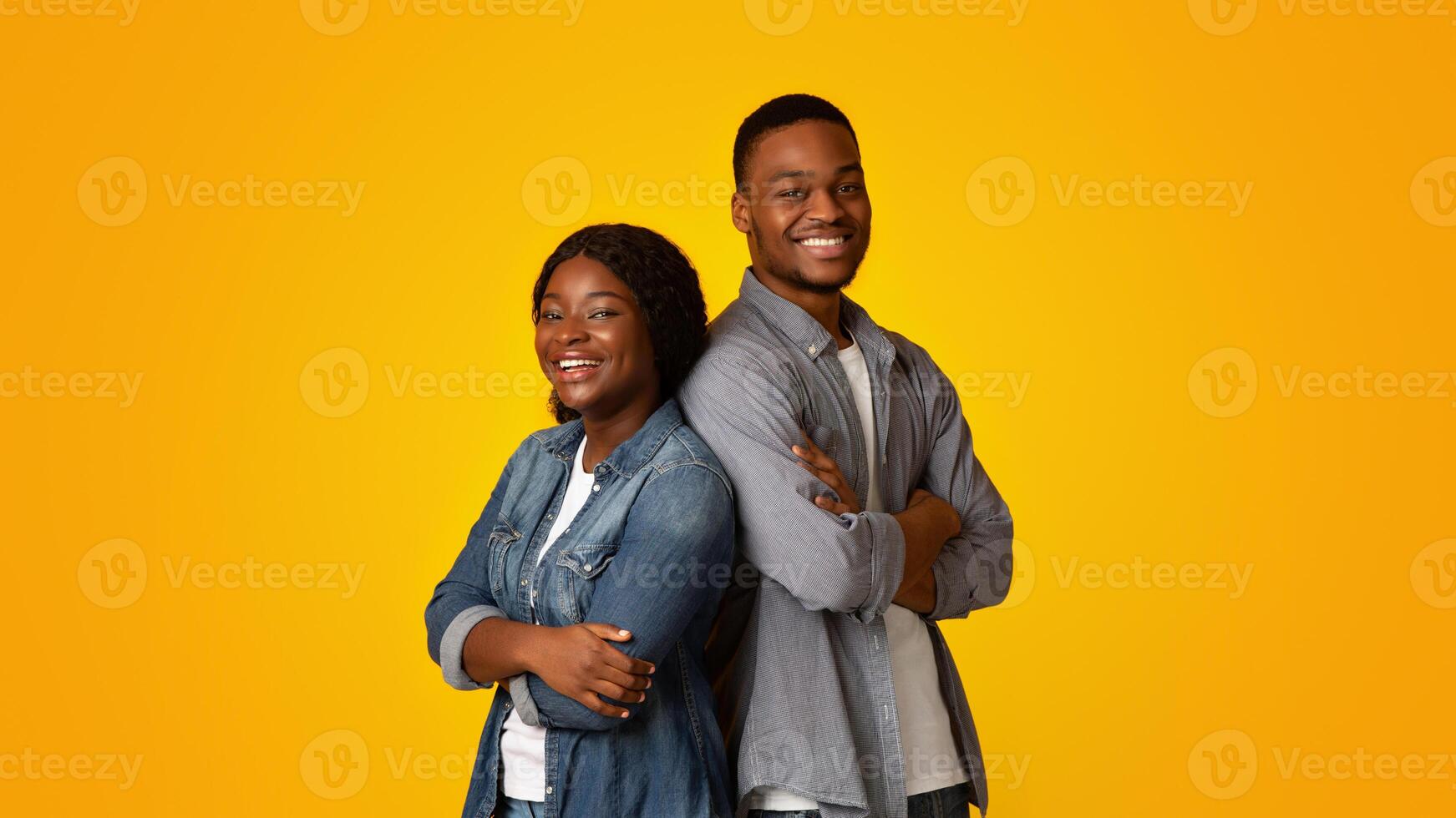 Portrait of confident happy young black man and woman standing back to back with folded arms, cheerful african american couple posing together over yellow background, looking and smiling at camera photo