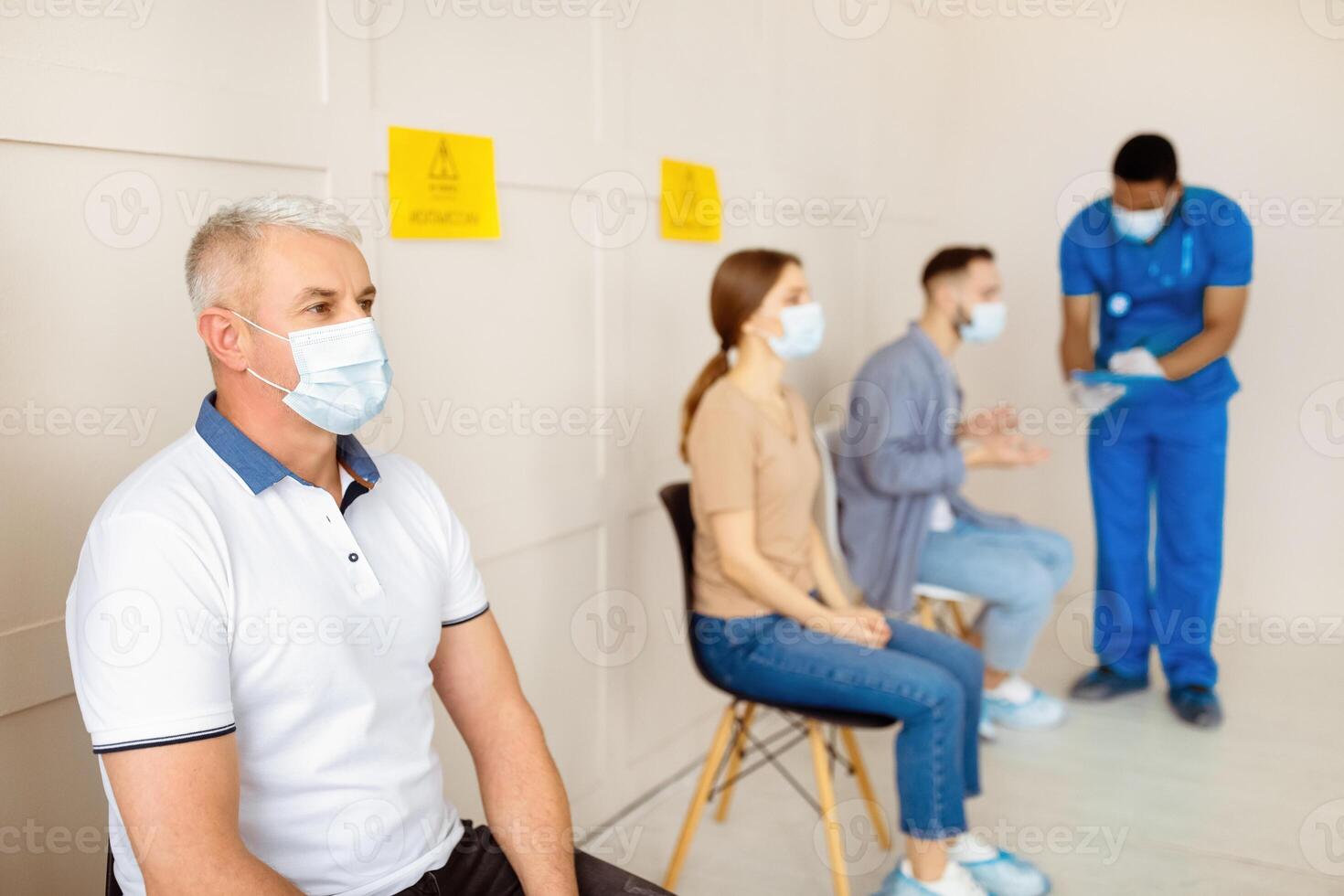 Senior man in protective mask sitting at hospital hall, waiting for covid-19 vaccine, nurse speaking to patients in queue. People participating in coronavirus immunization campaign. Healthcare concept photo