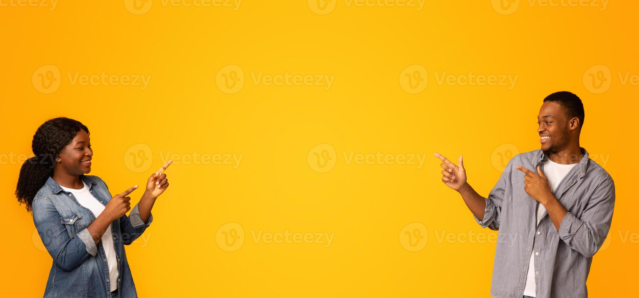 Look There. Joyful black couple pointing at copy space between them, demonstrating free place for your design or advertisement in the middle of yellow background, recommending something, panorama photo