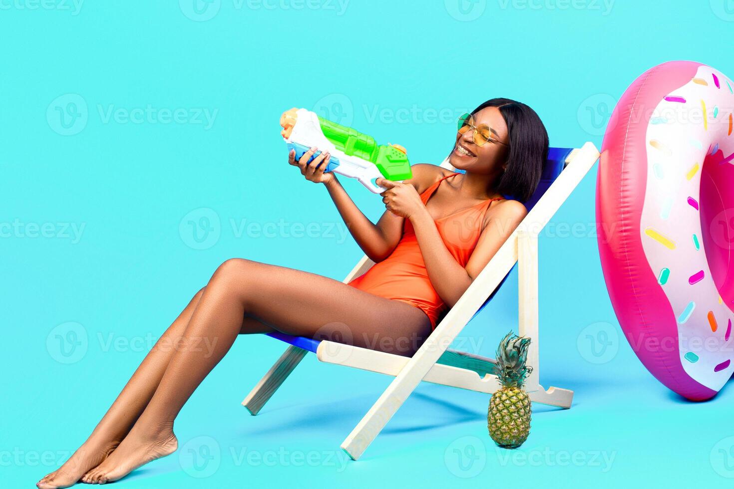 Summertime fun. Attractive black woman sitting in lounge chair with water gun, playing beach game over blue studio background, full length. Happy young lady chilling on tropical resort photo
