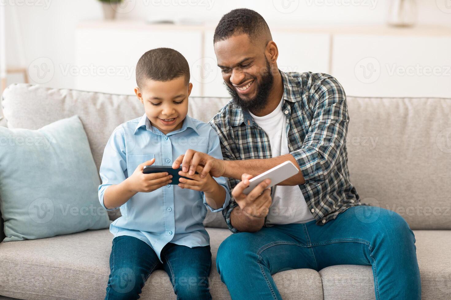 Black Father And Son Using Smartphones Sitting On Couch Together At Home. Mobile Games And Applications For Cellphone, Family And Modern Gadgets Lifestyle Concept. Selective Focus photo