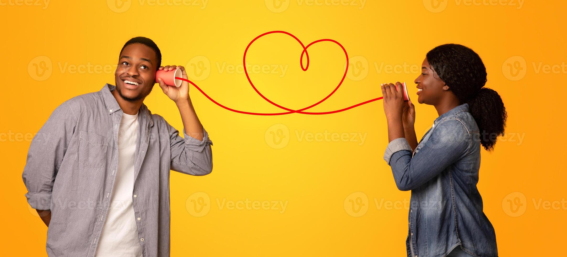 Love Message. Happy black woman whispering romantic words to her boyfriend via tin phone, standing isolated over yellow background, creative collage for Valentine's day with drawn red heart, panorama photo