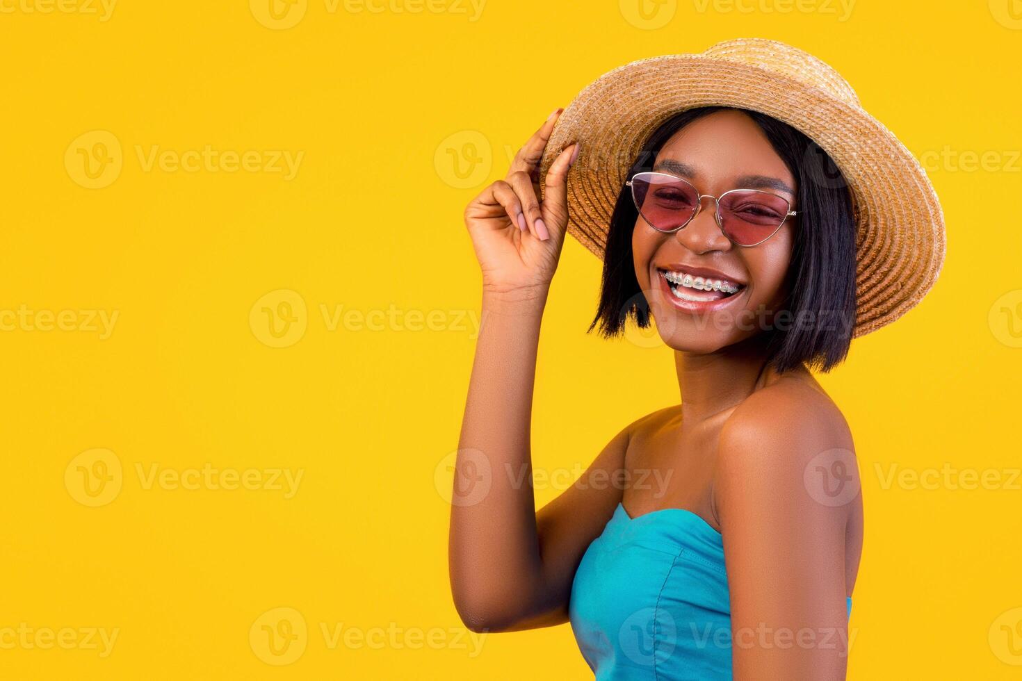 Smiling black lady in elegant dress and sunglasses touching her straw hat, ready for beach weekend on orange studio background. Posh African American woman posing in trendy summertime outfit photo