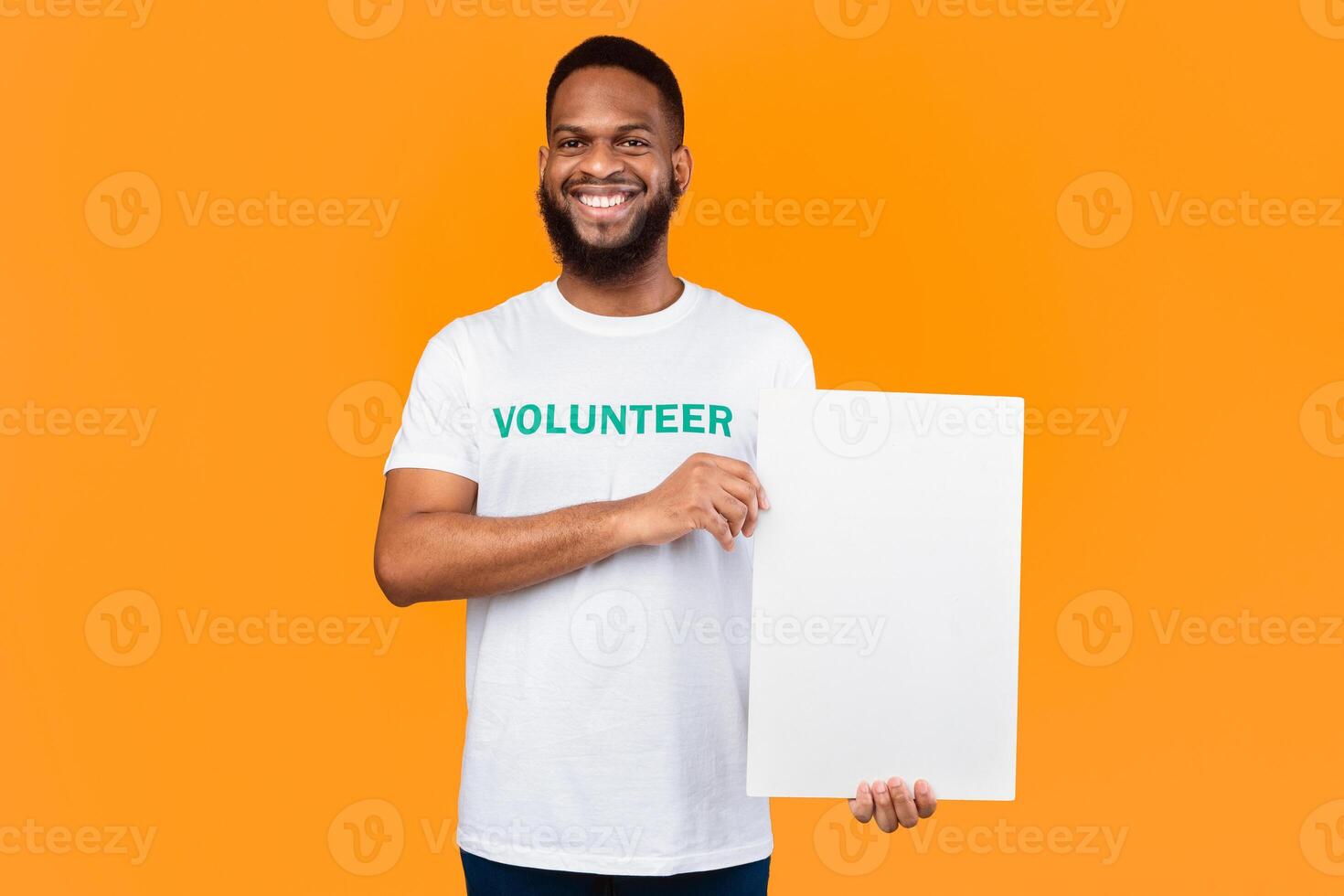 African American Volunteer Man Showing Blank White Paper Board Holding Empty Poster Advertising Your Text Posing Standing Over Yellow Background, Smiling To Camera. Studio Shot, Mockup photo