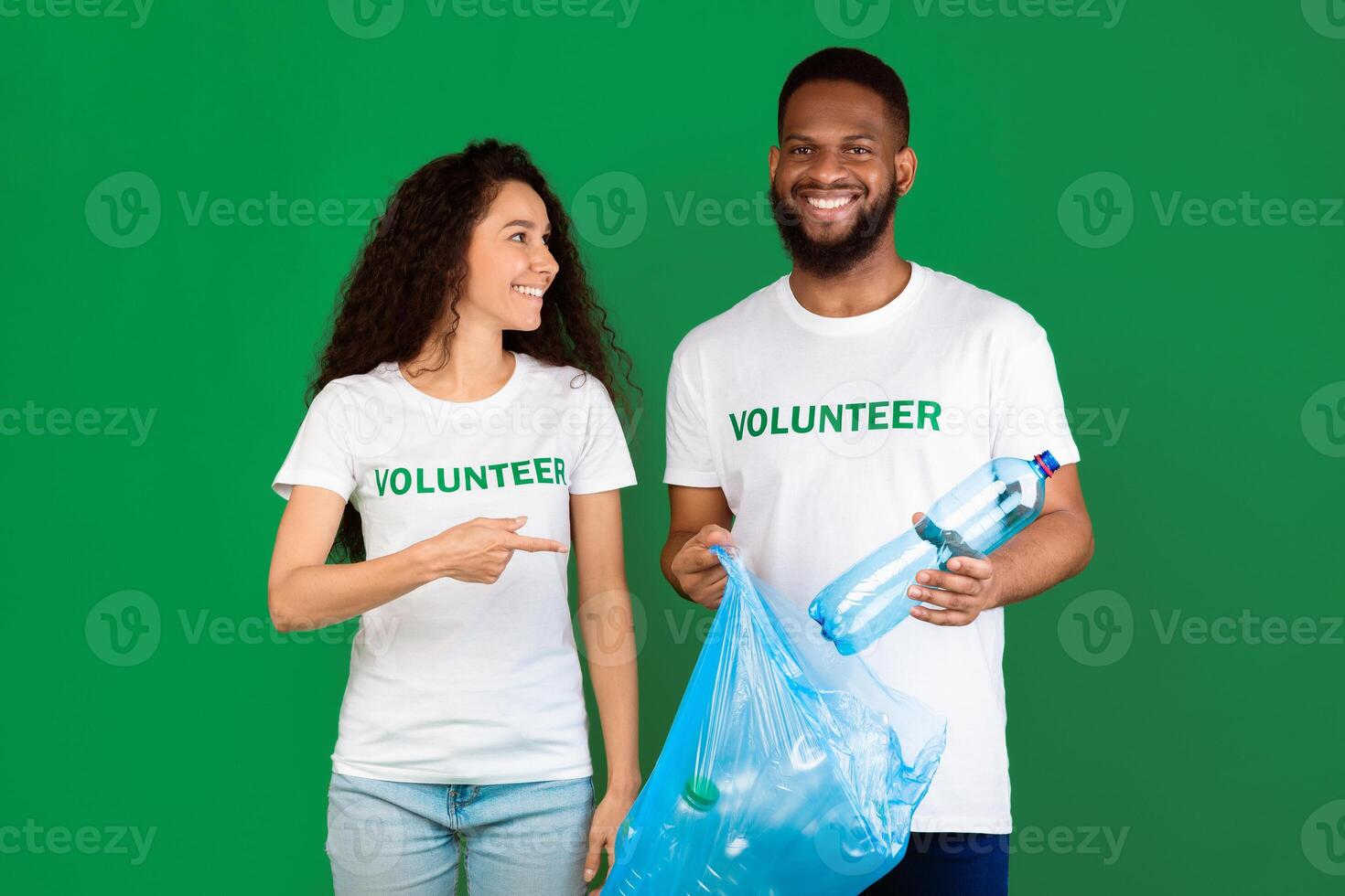 Two Diverse Volunteers Putting Wasted Plastic Bottle To Blue Garbage Bag Posing Standing On Green Background In Studio. Junk Sorting, Ecology And Volunteering Work, Keep Environment Clean Concept photo