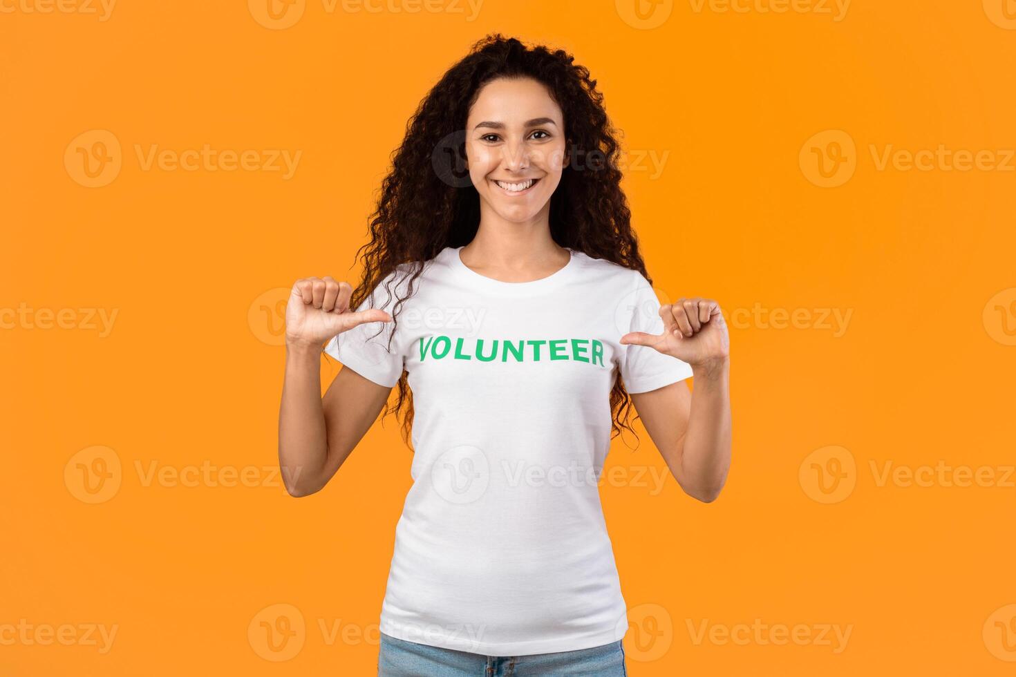 I'm a volunteer. Happy woman activist pointing at herself and volunteer inscription on her t-shirt standing over yellow studio background, smiling to camera. Volunteering Job Concept photo