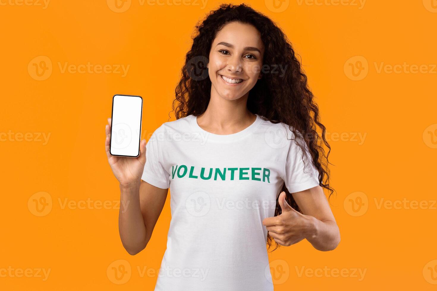 Happy Volunteer Lady Showing Blank Smartphone Screen And Gesturing Thumbs-Up To Camera Posing Standing Over Yellow Studio Background. Mobile App For Charity And Volunteering Concept. Mockup photo