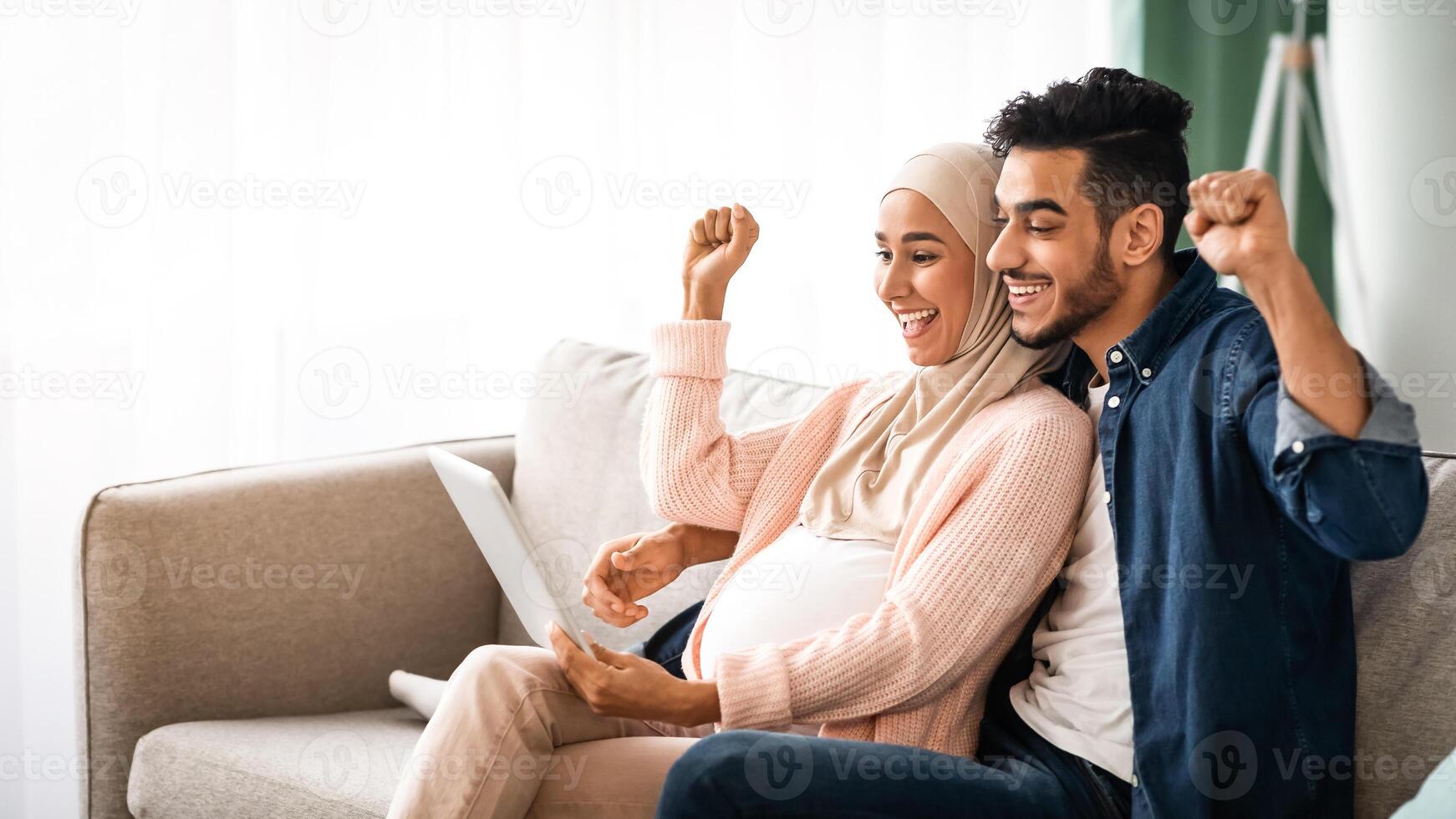 Excited Pregnant Muslim Couple Celebrating Success With Digital Tablet At Home, Happy Expectant Middle Eastern Spouses Looking At Tab Computer Screen, Shaking Fists And Exclaiming With Excitement photo