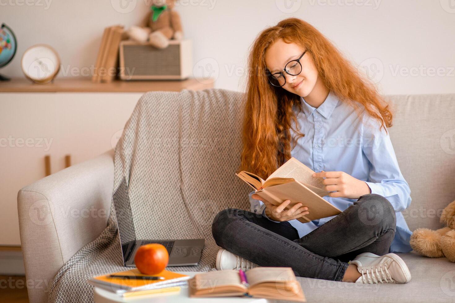 Leisure, Relaxation And Cozy Weekend. Smiling female teenager in eyewear holding and reading paper book. Schoolgirl sitting on the comfortable couch with laptop at home in bedroom, taking a break photo
