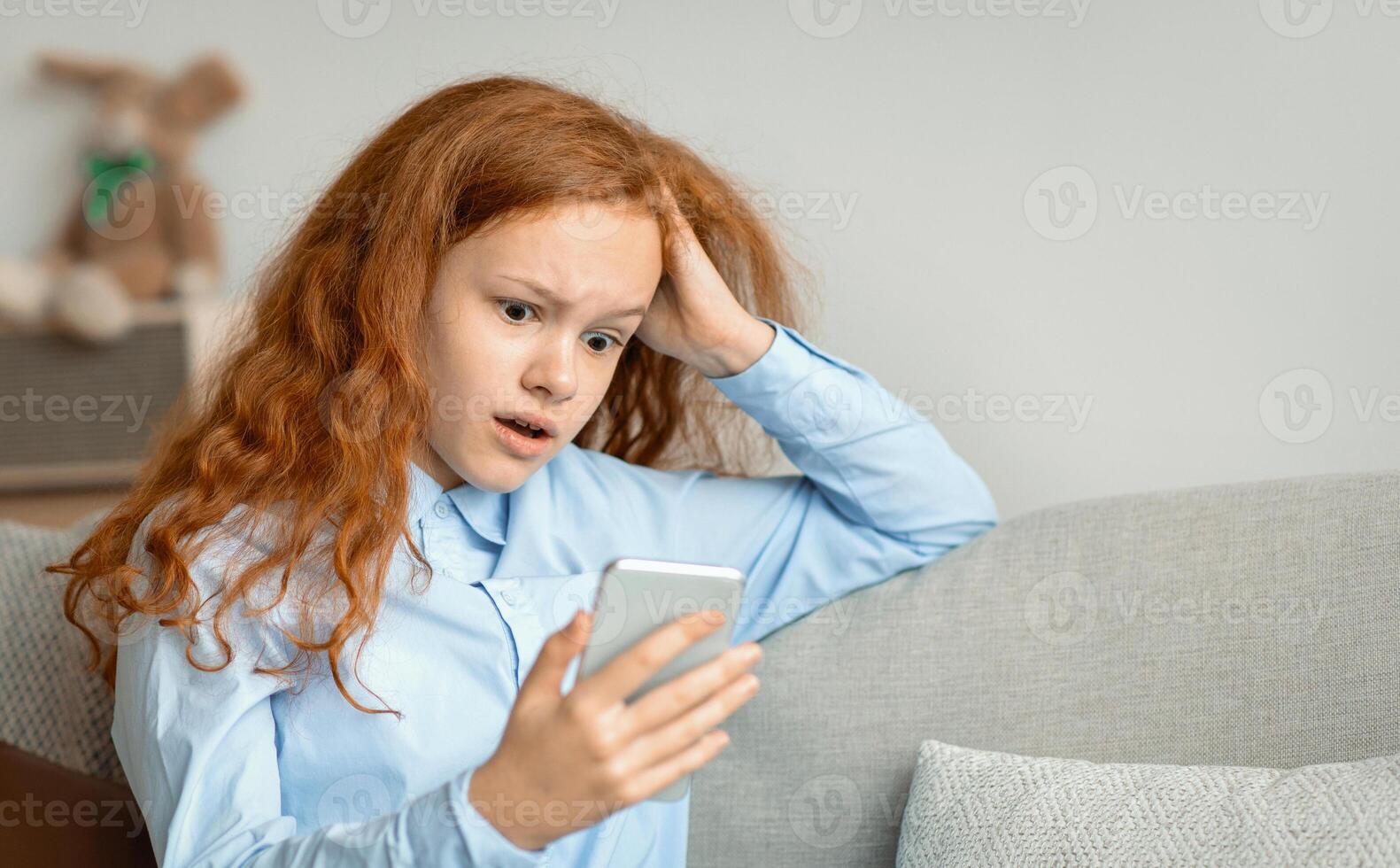 Shocked Surprised Young Teen Girl With Cellphone Reading Bad News Message Touching Head In Amazement Sitting On The Couch At Home. Confused Mobile Phone User, Call From Unknown Number photo