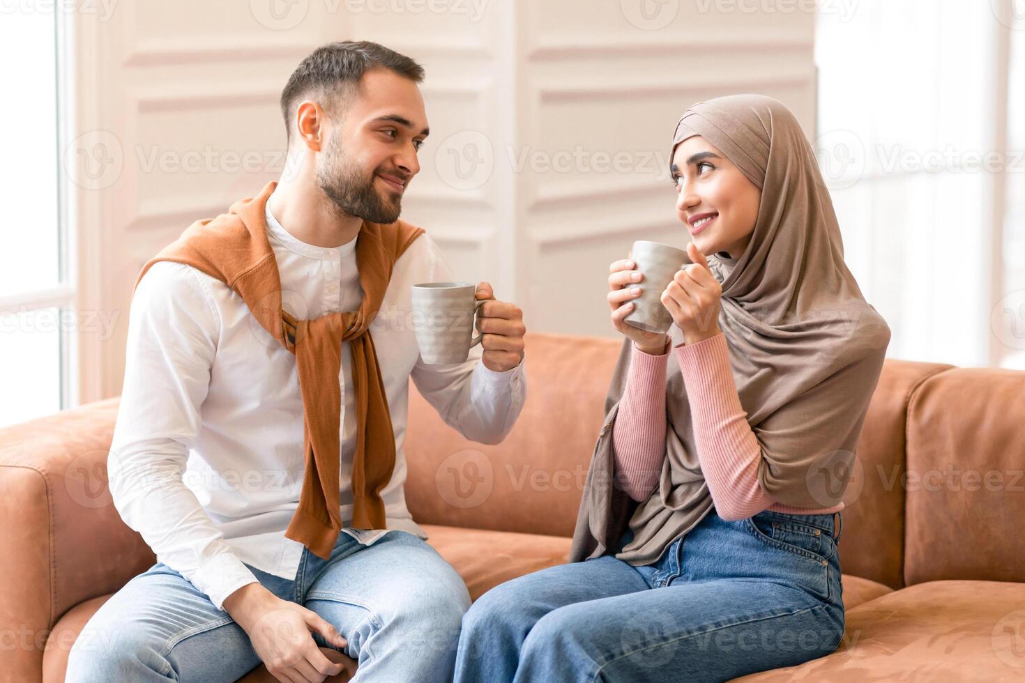 Romantic Relationship. Cheerful Muslim Spouses Drinking Coffee Sitting On Couch Smiling Each Other Posing At Home. Arabic Young Couple Flirting And Bonding Spending Time Together On Weekend Indoors. photo