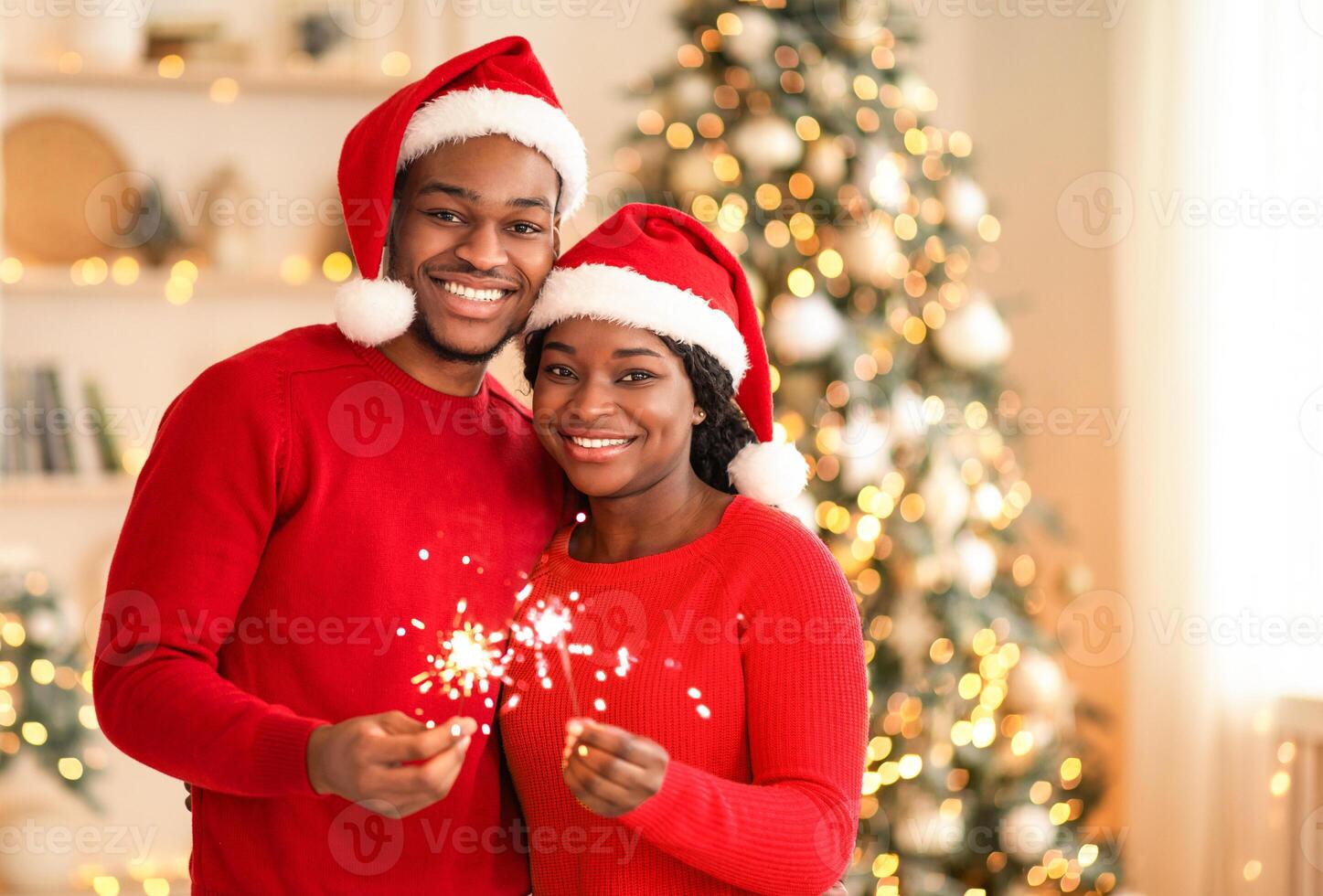 Happy New Year and Merry Xmas at home together. Smiling young african american guy and lady in Santa Claus hats hold sparklers, hug and look at camera in living room interior with tree and garlands photo