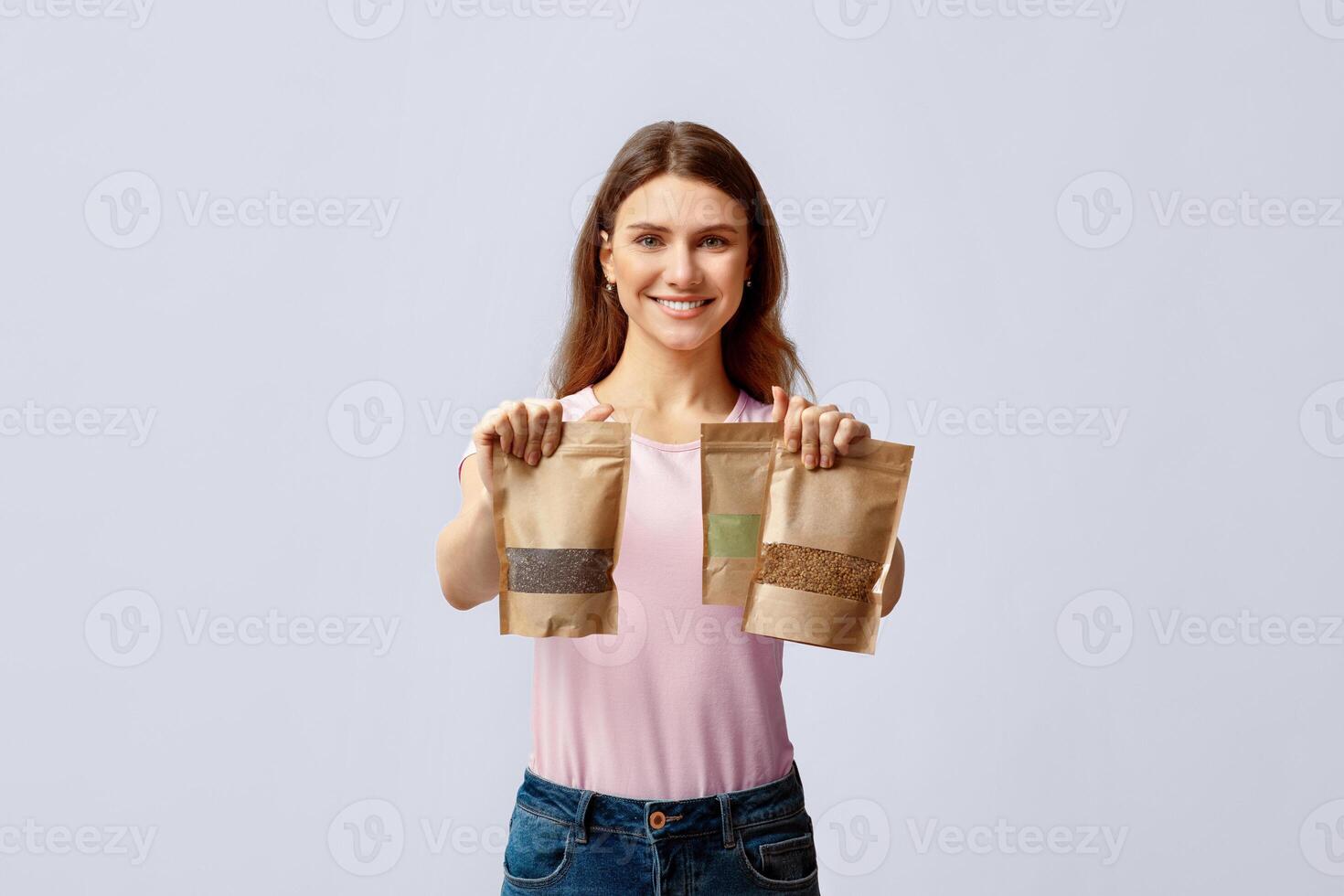 Eco Food Storing. Positive Young Lady Holding Three Kraft Paper Doypack Bags With Weight Groceries, Smiling Millennial Woman Choosing Biodegradable Ziplock Packs, Posing Over Light Grey Background photo