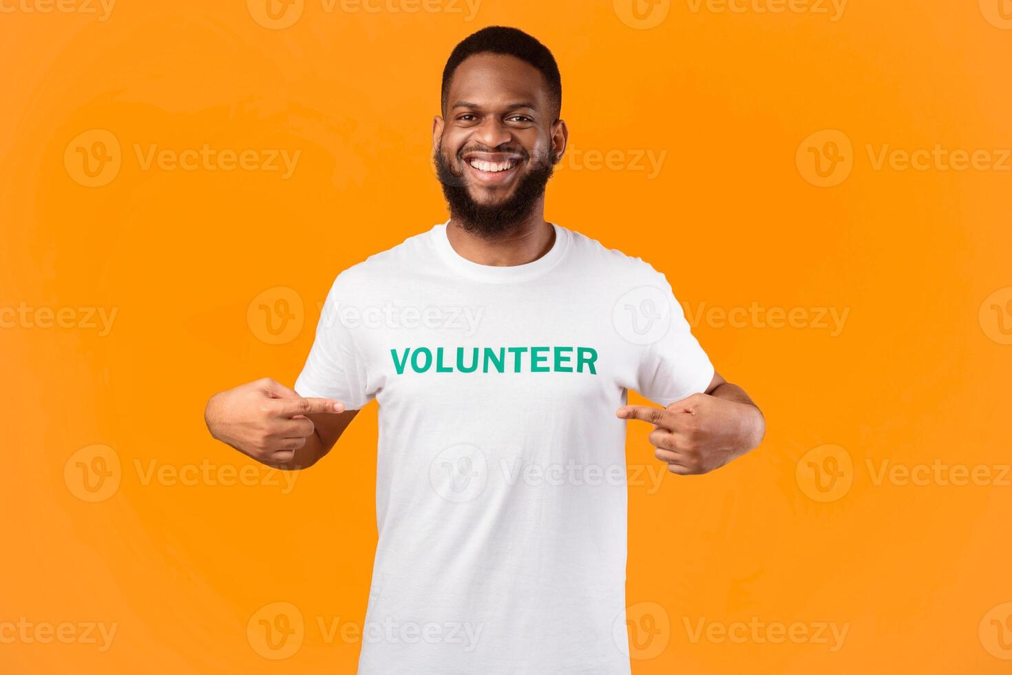 Happy african american volunteer guy showing his uniform t-shirt with text, smiling to camera posing on yellow background, studio shot. Portrait of joyful activist. I choose volunteering career photo