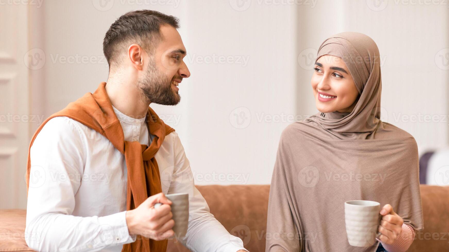 Young Muslim Couple Talking And Flirting Drinking Coffee Enjoying Conversation Sitting On Couch Smiling Each Other At Home. Beautiful Arab Woman Touching Husband's Hand. Love, Romantic Relationship photo
