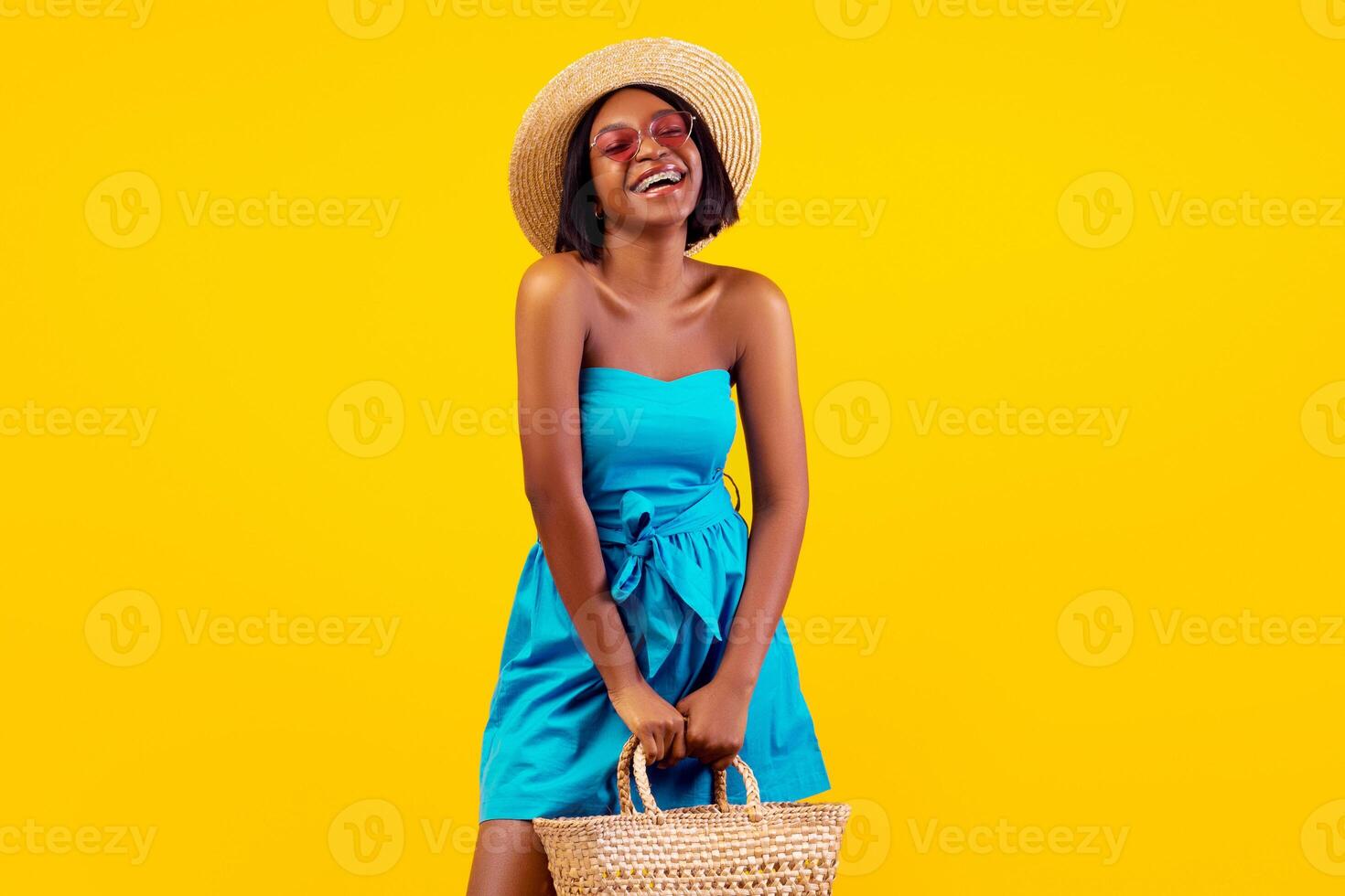 Beautiful black woman in elegant summer dress and straw hat holding beach bag, laughing and enjoying her vacation on orange studio background. Summertime holiday or weekend concept photo