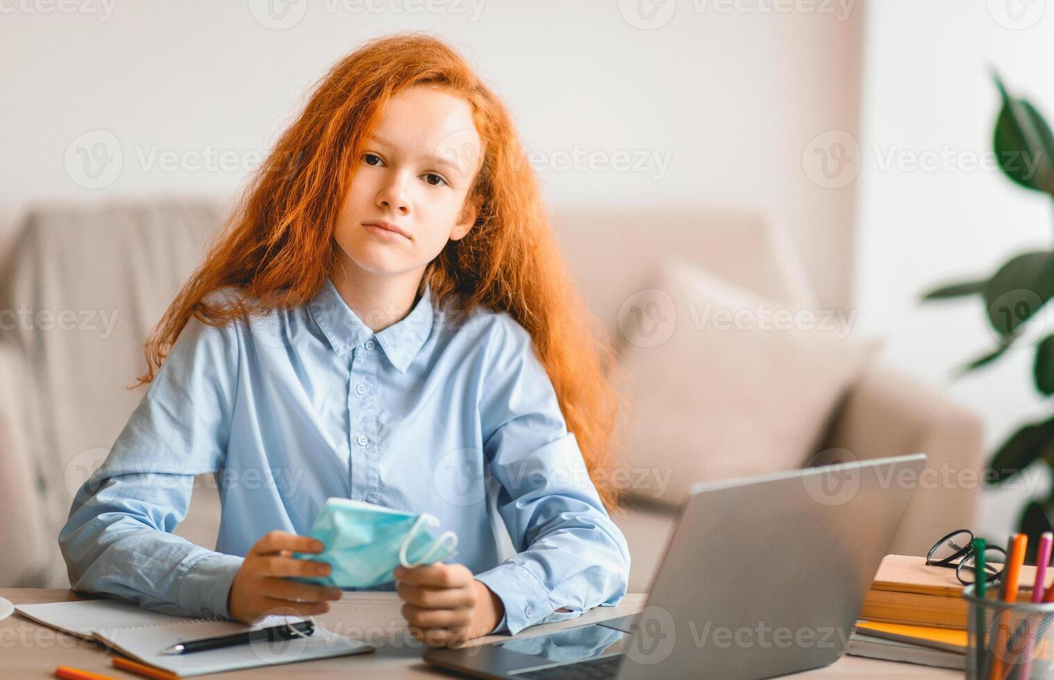 Quarantine And Lockdown Concept. Portrait of sad teenage schoolgirl holding disposable medical face mask, studying and learning at home using her laptop computer. Upset bored girl looking at camera photo