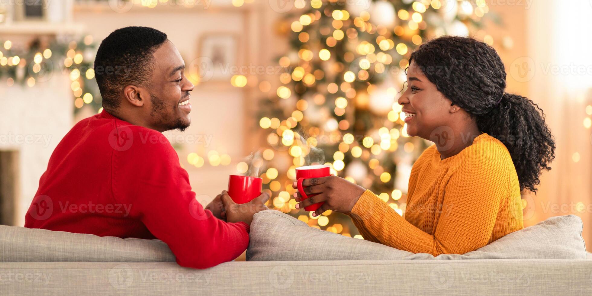 Favorite drink and New Years mood at home. Happy millennial african american family with cups of cocoa look at each other in interior of home with tree and glowing garlands, profile, copy space photo
