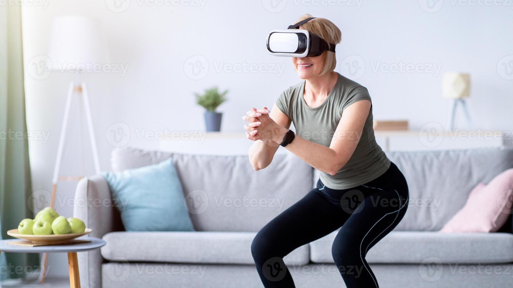 Exercising with virtual reality concept. Athletic mature woman in VR headset doing squats on her home workout. Positive senior lady using modern technologies for domestic training photo