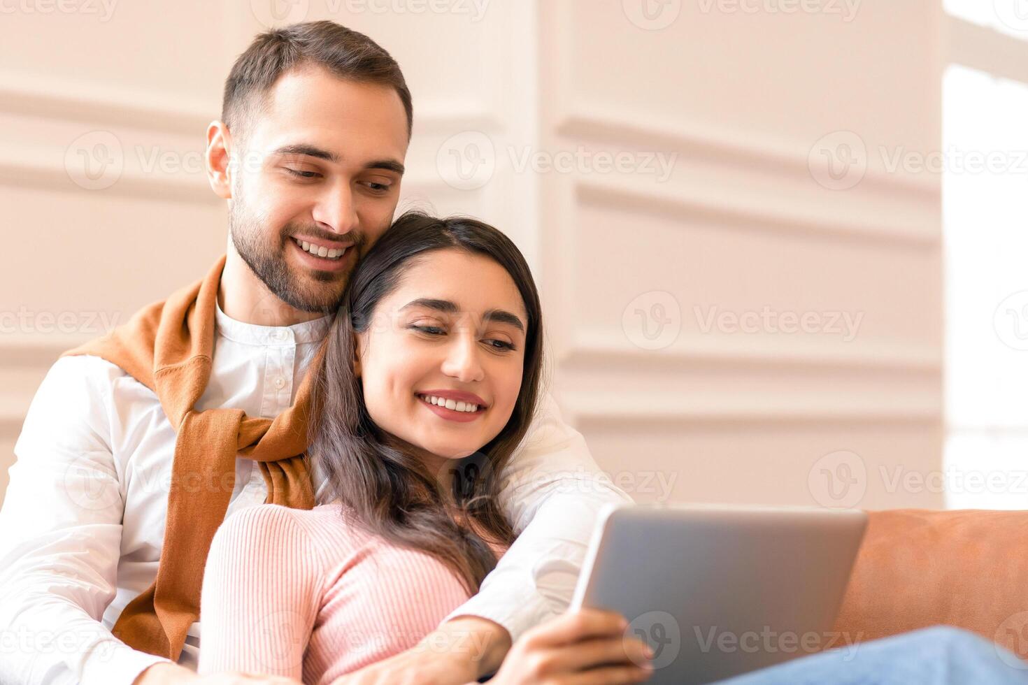Happy Modern Arab Couple Using Digital Tablet Computer Watching Movie Online Sitting On Sofa At Home. Spouses Browsing Internet Together Indoors. Family Weekend Concept photo