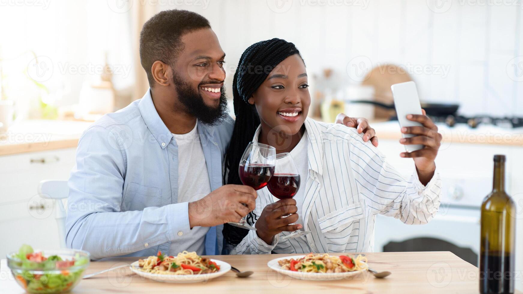 Happy Moments. Cheerful Black Couple Taking Selfie On Smartphone While Having Romantic Date At Home, Young African American Spouses Sitting At Table In Kitchen And Clinking Glasses With Red Wine photo