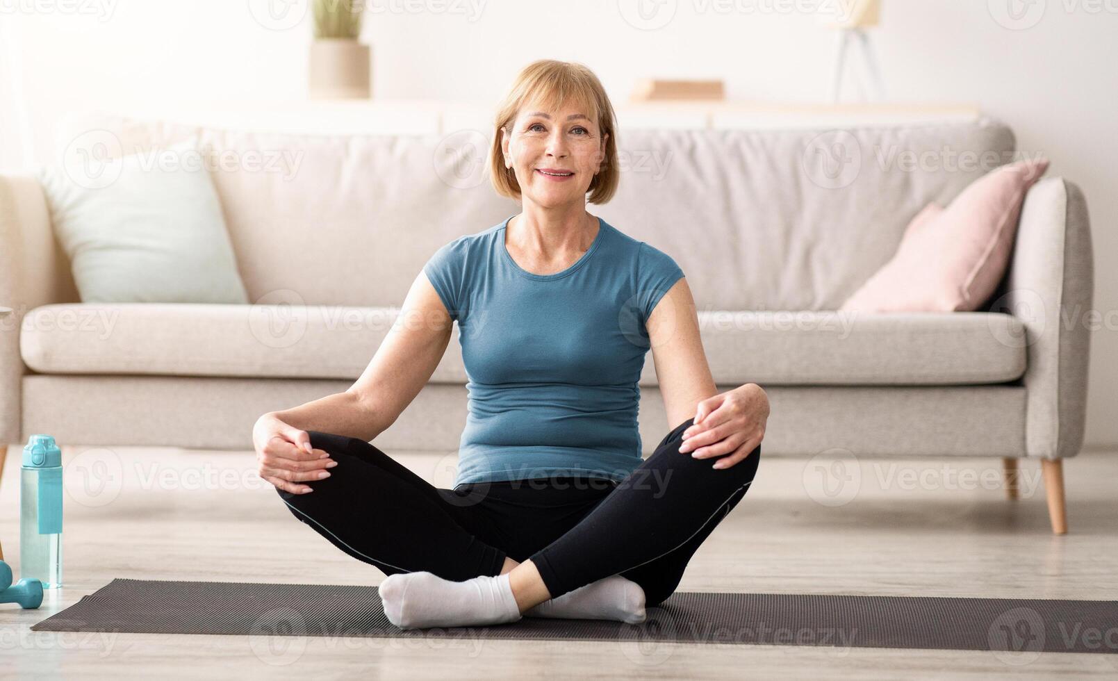 Positive senior woman sitting in lotus pose on sports mat, meditating at home, copy space. Balanced mature lady practicing yoga, finding inner peace and mindfulness photo