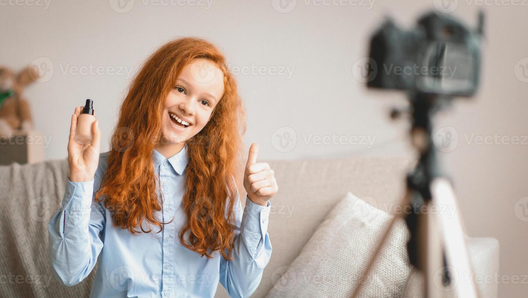 Beauty Advice And Blog. Smiling female teen doing makeup, recording cosmetics product review, showing her foundation for young skin to camera on tripod. Girl filming , showing thumbs up photo
